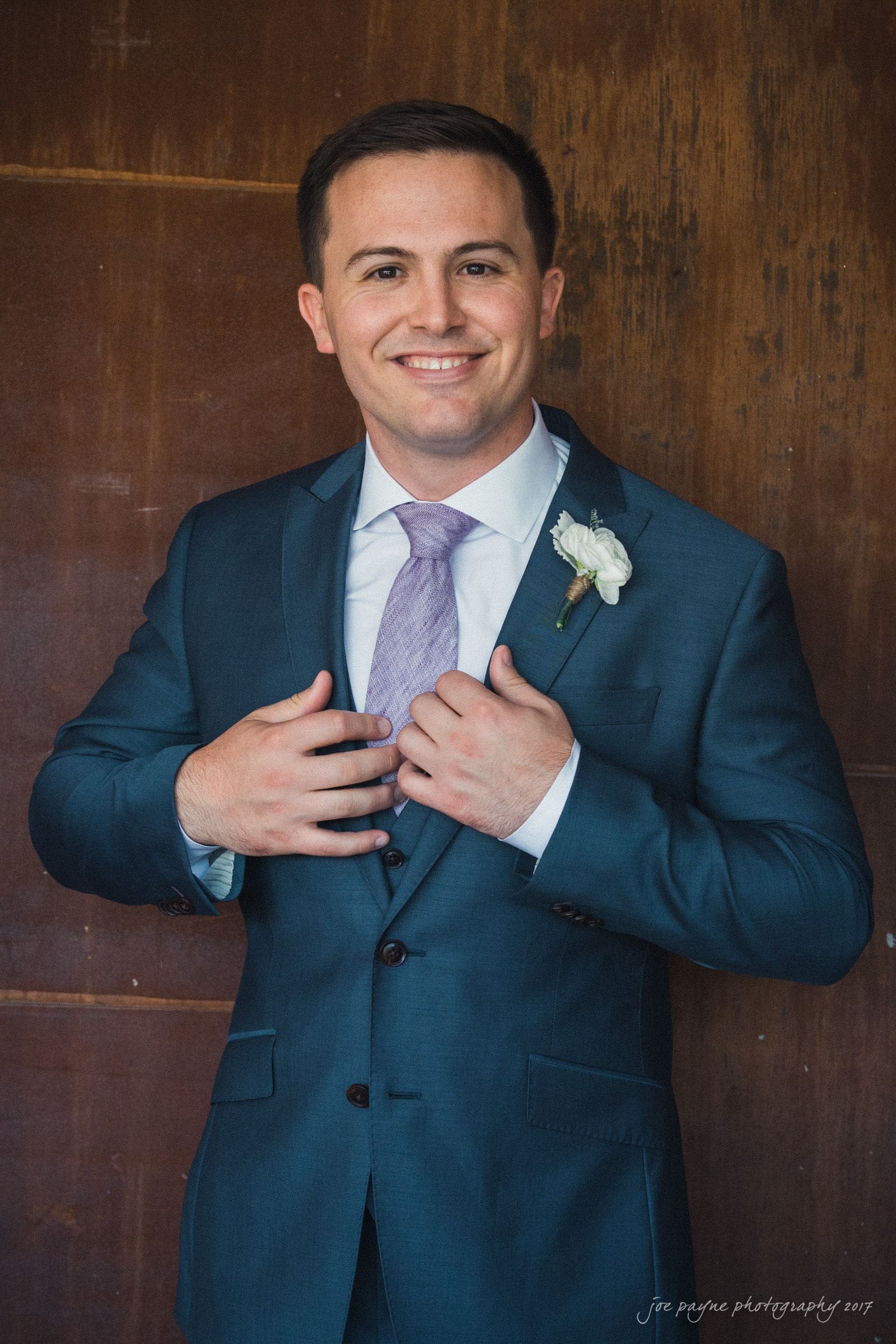 melrose knitting mill wedding - natalie & elliot A smiling man in a tailored blue suit and lavender tie adjusts his jacket. He has short dark hair and a boutonniere on his lapel, standing against a rustic brown background.