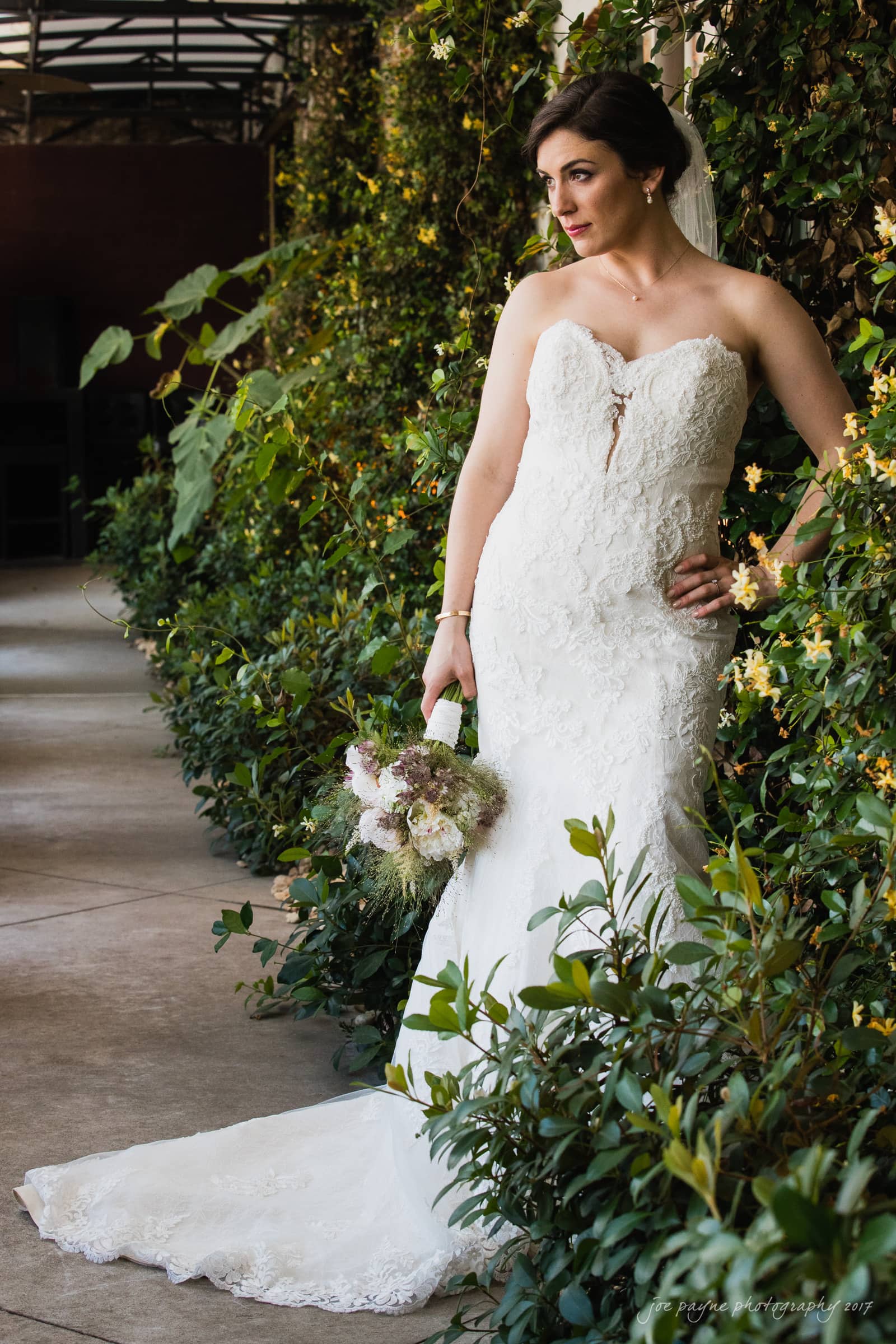 melrose knitting mill wedding - natalie & elliot A bride in a strapless lace gown poses against a greenery-covered wall, holding a bouquet of flowers. She looks thoughtfully into the distance, with one hand on her hip and the other holding the bouquet at her side.