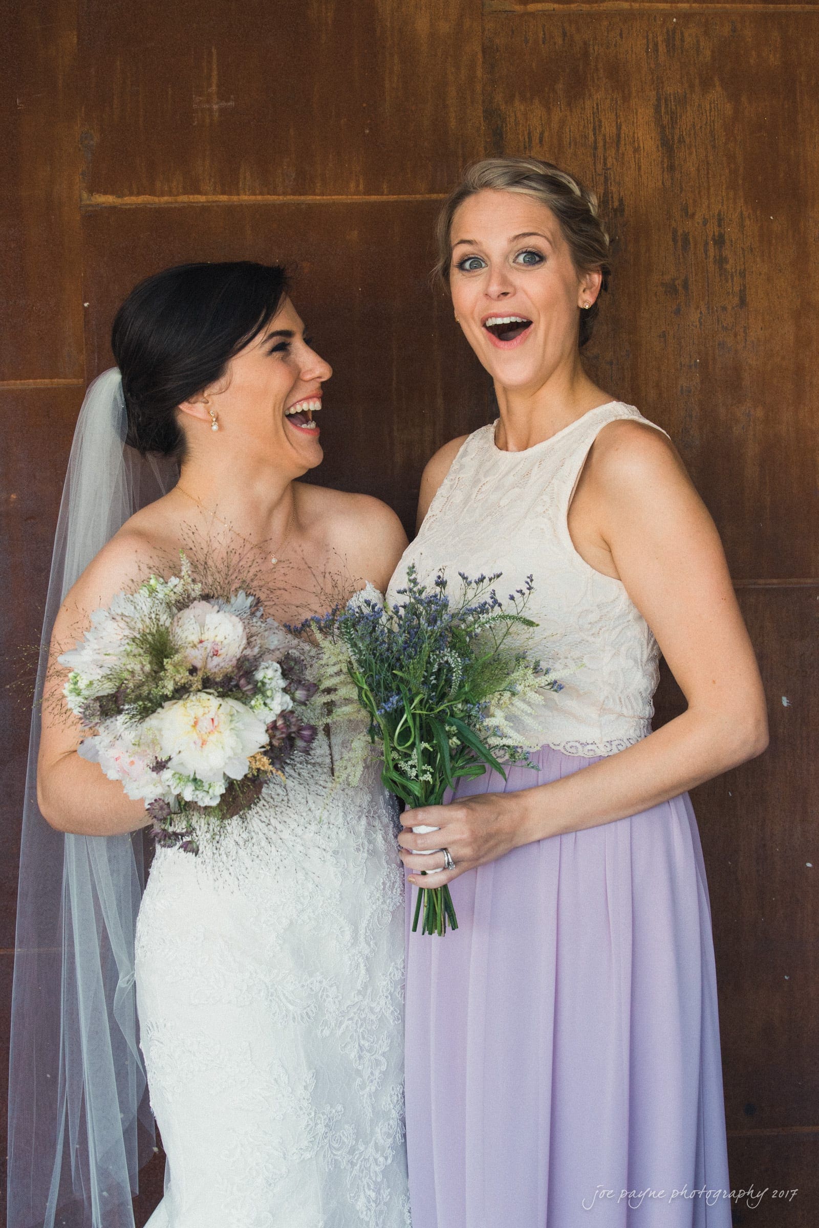 melrose knitting mill wedding - natalie & elliot A bride and a bridesmaid stand next to each other, both smiling and holding bouquets. The bride wears a white strapless dress and veil, while the bridesmaid wears a lavender dress. The background is a rustic brown wall.