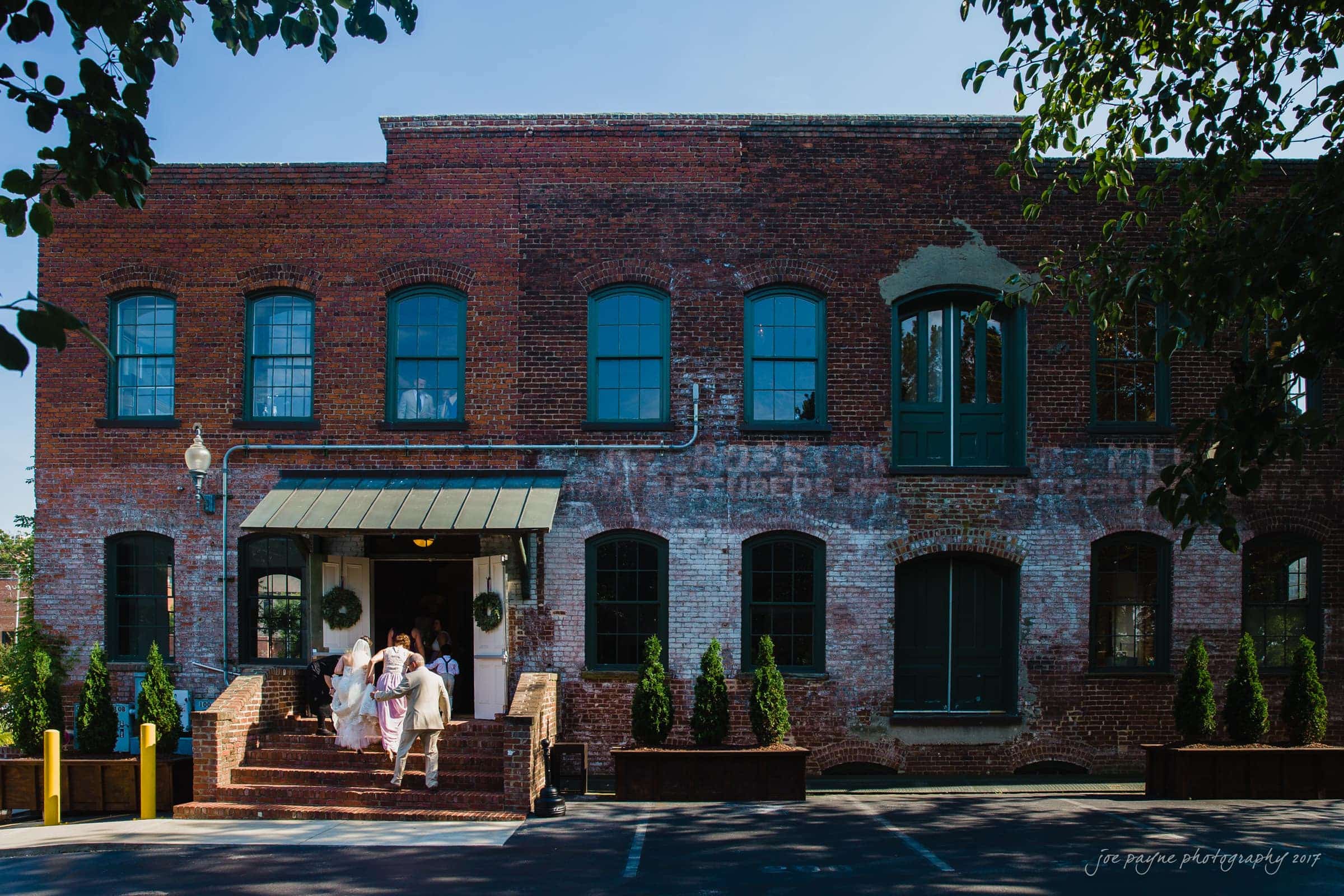 melrose knitting mill wedding - natalie & elliot A group of people dressed formally stand on the steps of a rustic, red-brick building with large green doors and windows. The scene is set on a sunny day, and there are trees framing the image.