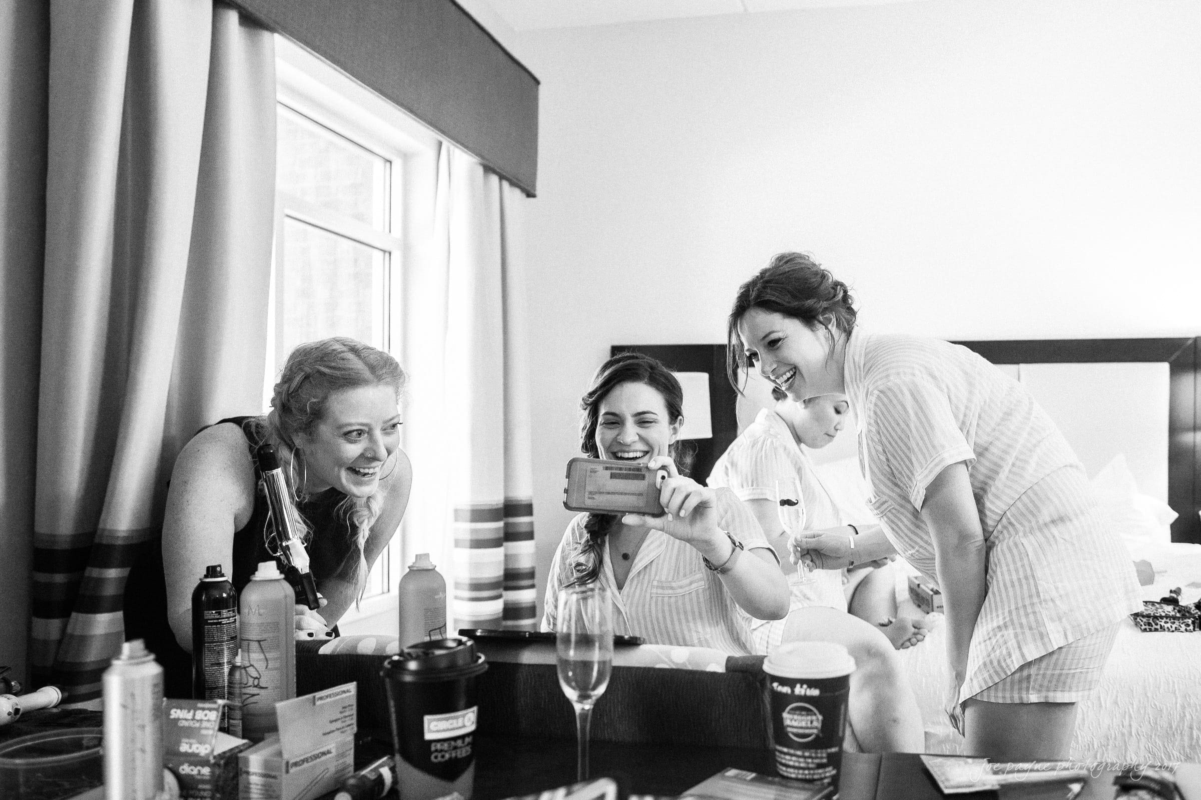melrose knitting mill wedding - natalie & elliot A group of three women and a baby enjoy a moment together in a room. The women are looking at a smartphone, smiling and laughing. The setting includes a table with various items like a coffee cup and a champagne glass.