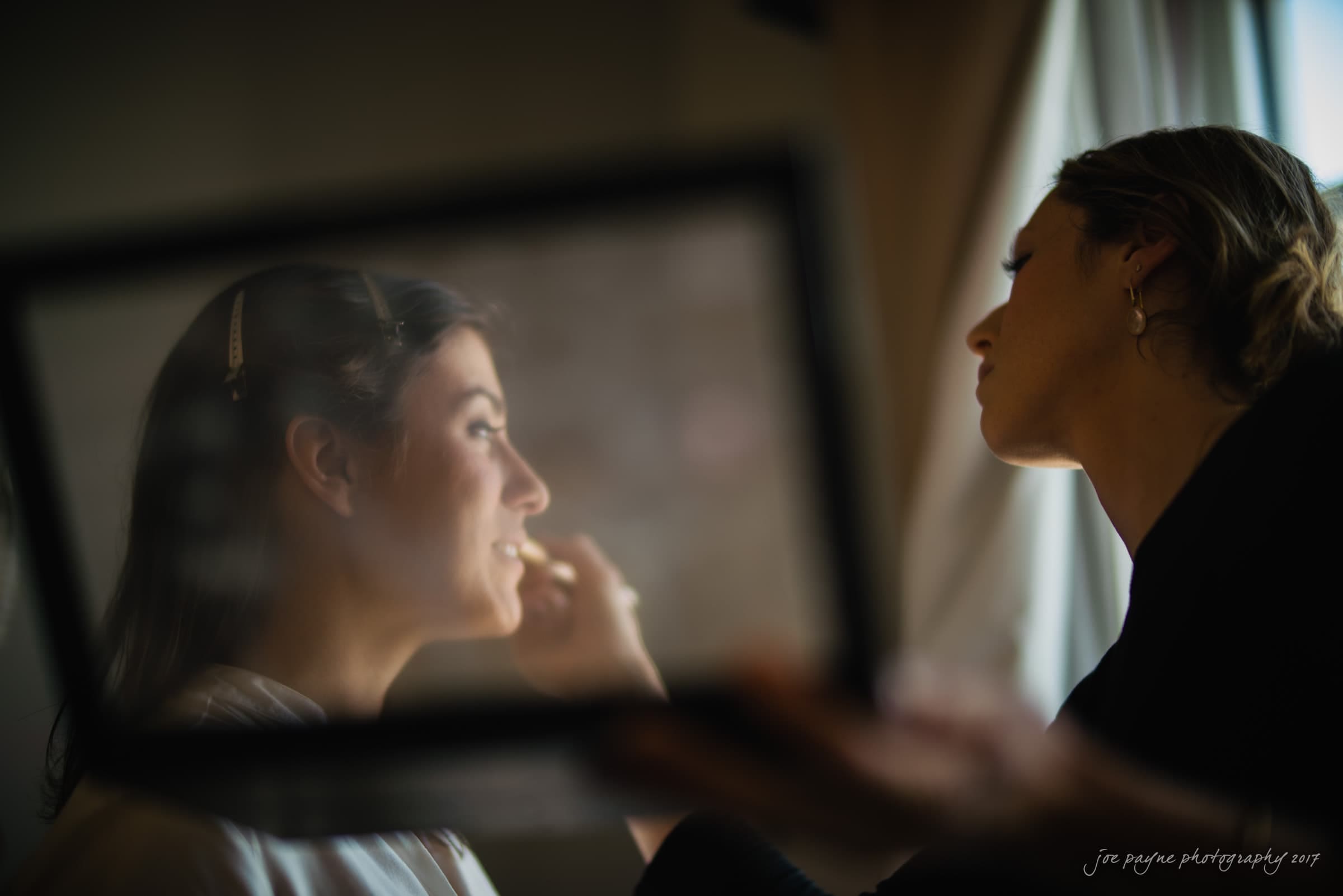 melrose knitting mill wedding - natalie & elliot A bride is getting her makeup done by a makeup artist. The artist is applying lipstick, and the bride is smiling while looking into a mirror. The scene is softly lit, creating a serene atmosphere.