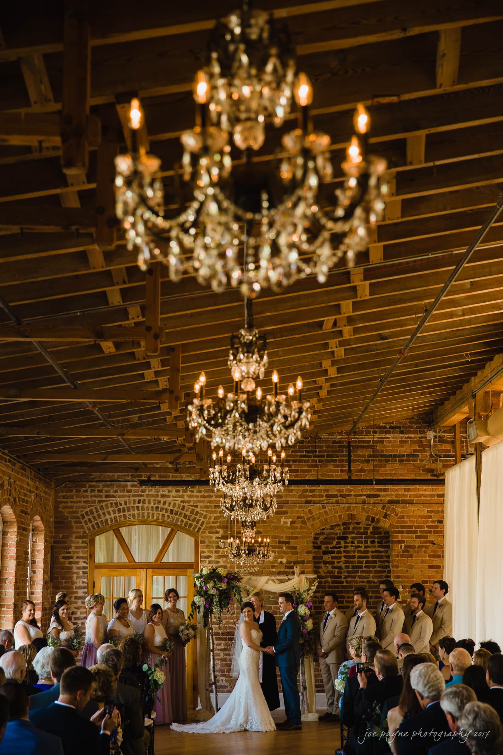 melrose knitting mill wedding - natalie & elliot A wedding ceremony in a rustic venue with exposed brick walls and overhead chandeliers. The couple stands at the altar, surrounded by the bridal party and seated guests. The space is warmly lit, highlighting the intimate and elegant atmosphere.