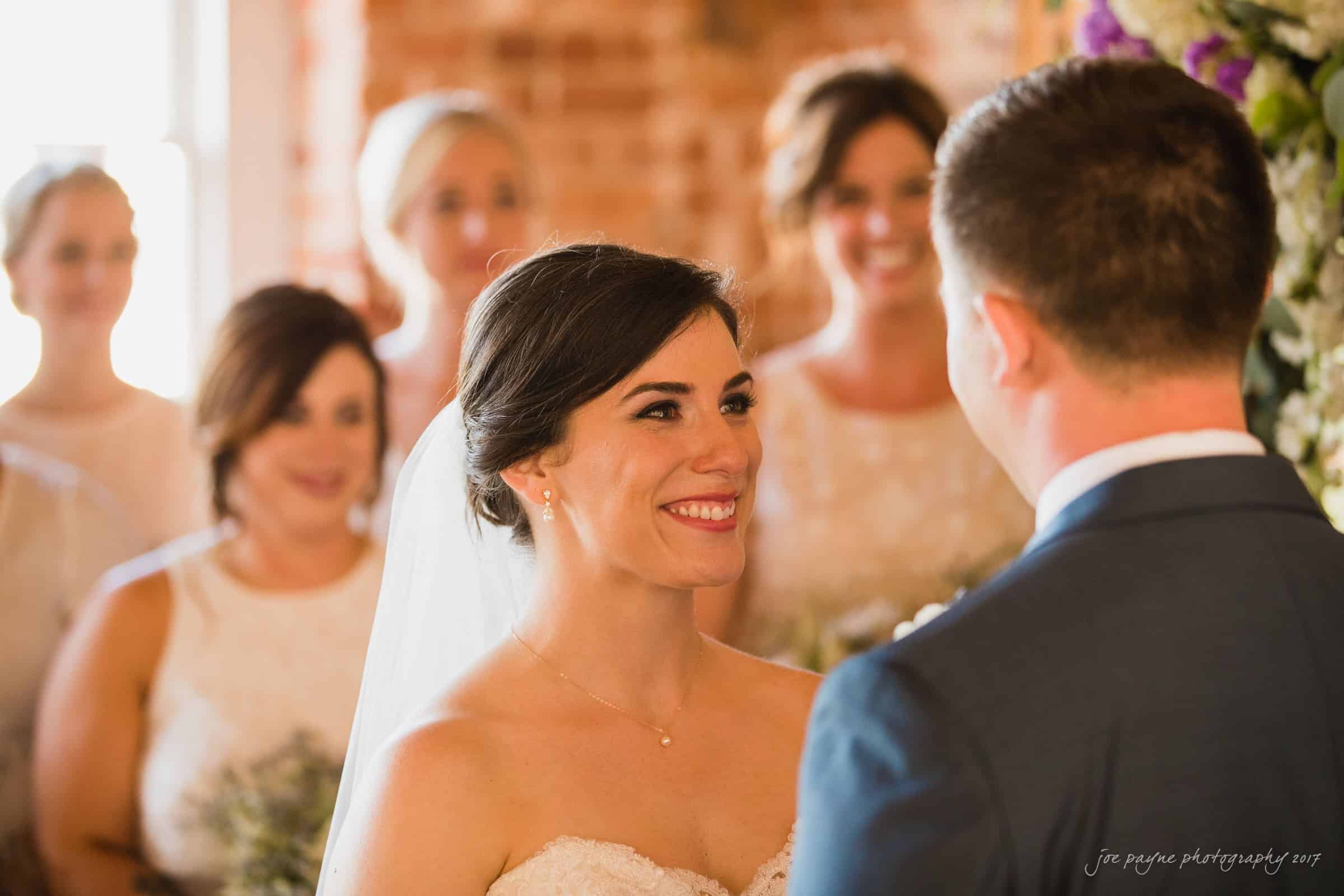 melrose knitting mill wedding - natalie & elliot A bride smiles warmly at her groom during a wedding ceremony. Bridesmaids in light dresses stand in the soft-focus background. The setting has rustic brick walls and floral decor.