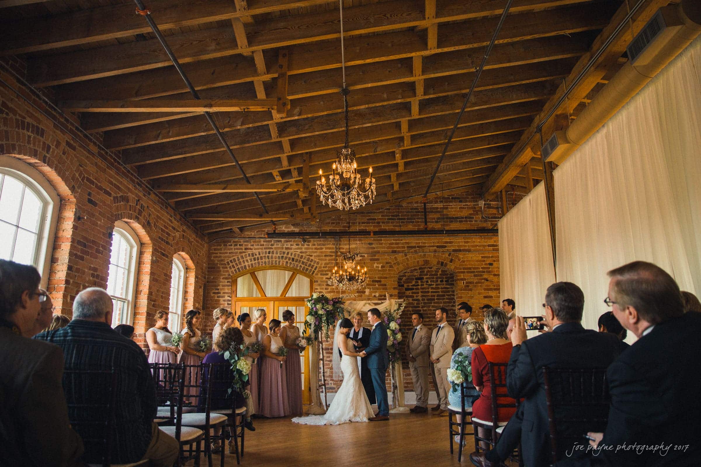 melrose knitting mill wedding - natalie & elliot A wedding ceremony set in a rustic venue with brick walls and chandeliers. The couple stands at the altar surrounded by their bridal party. Guests are seated on either side, some capturing the moment with phones and cameras.