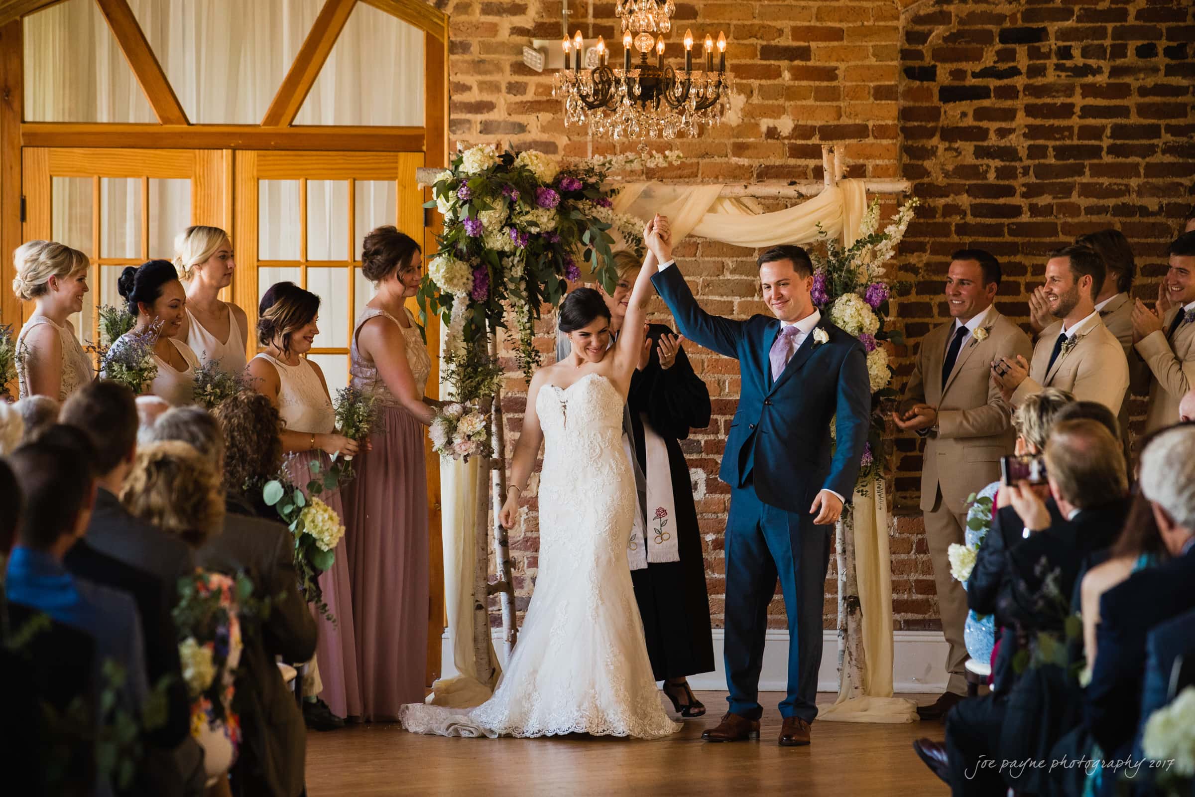 melrose knitting mill wedding - natalie & elliot A joyful couple stands at the altar, raising their hands in celebration during a wedding ceremony. The bride wears a white gown, and the groom is in a dark suit. Bridesmaids and groomsmen smile and clap. The backdrop features flowers and a chandelier.