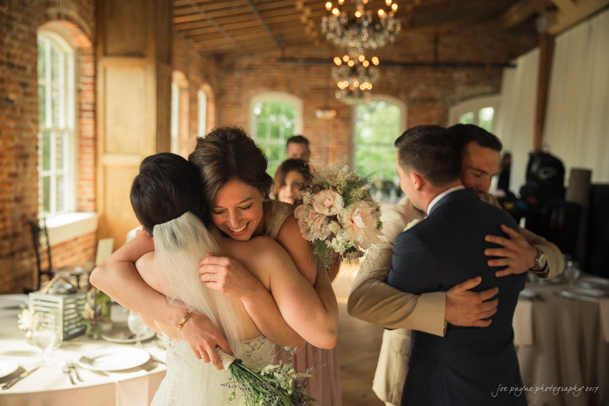 melrose knitting mill wedding - natalie & elliot A joyful wedding scene where two women in dresses share a warm embrace, one holding a bouquet. Nearby, two men in suits hug. The room has brick walls, large windows, and chandeliers, creating a cozy atmosphere.