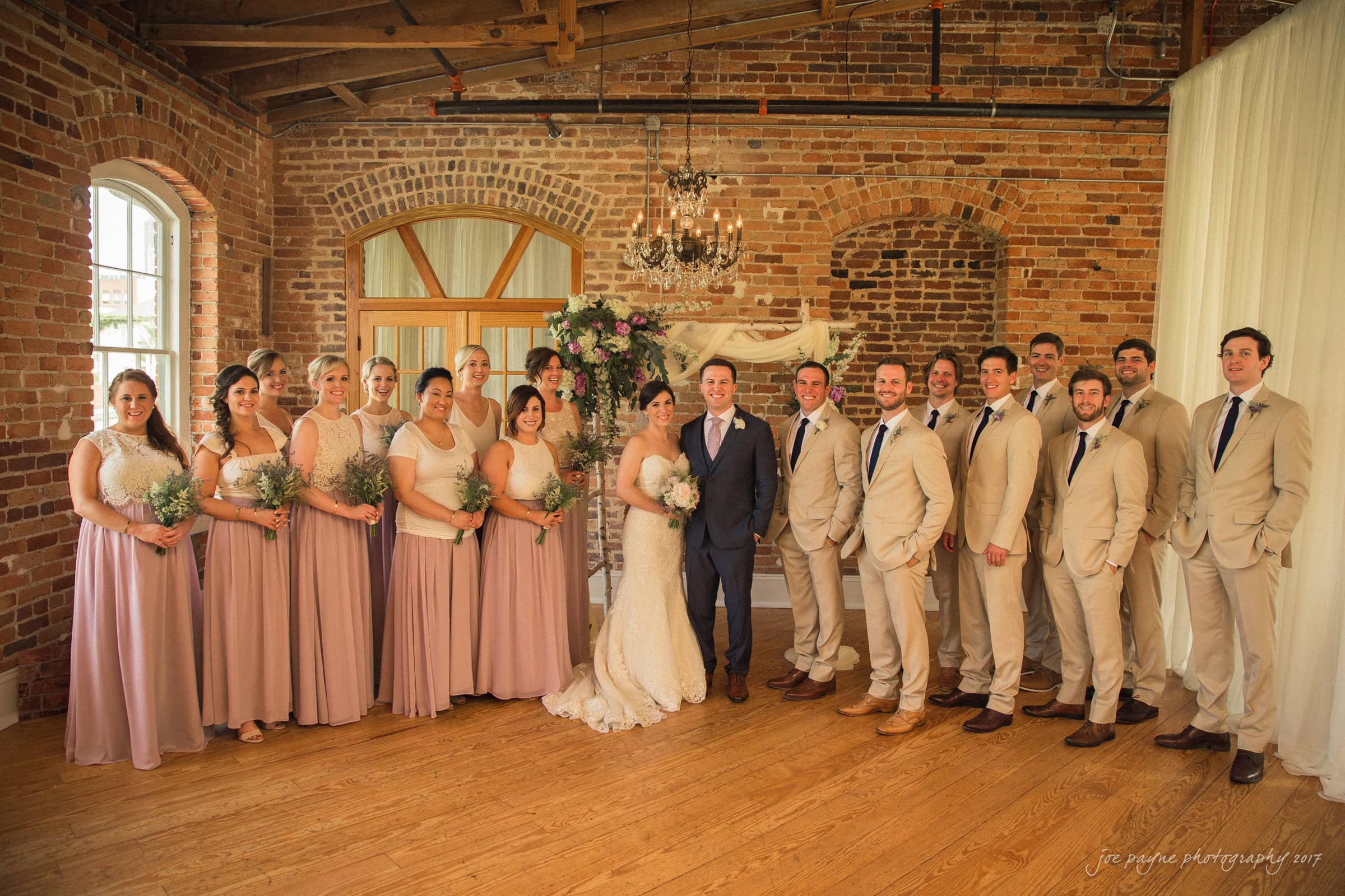 melrose knitting mill wedding - natalie & elliot A wedding party poses indoors against a brick wall. The group includes the bride and groom at the center, with bridesmaids in pink dresses and groomsmen in beige suits standing on either side. Floral arrangements are visible in the background.
