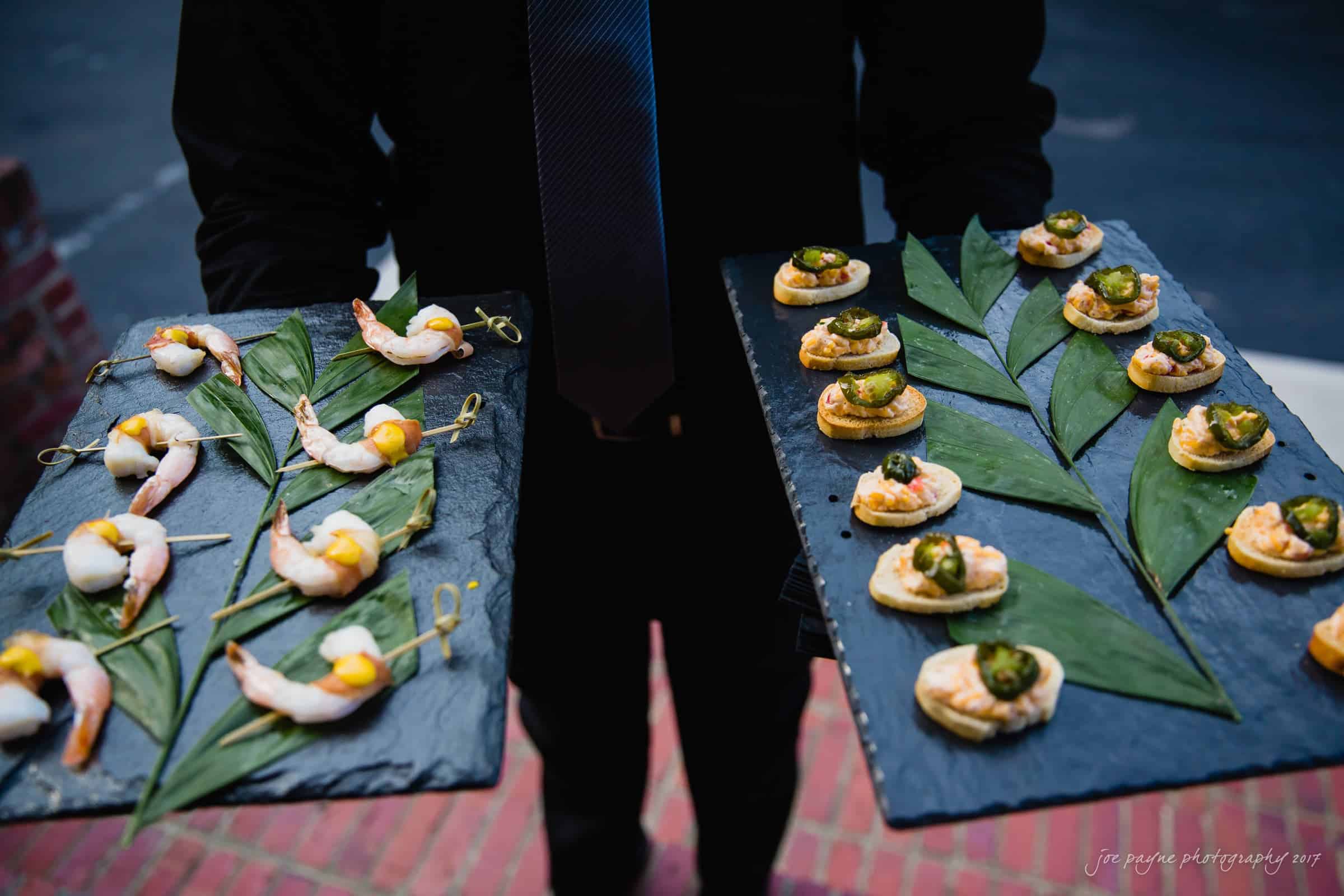 melrose knitting mill wedding - natalie & elliot A server holds two slate trays filled with appetizers. The tray on the left features skewered shrimp with a yellow garnish on leaves, and the tray on the right has small, round pastries topped with green and black garnishes.