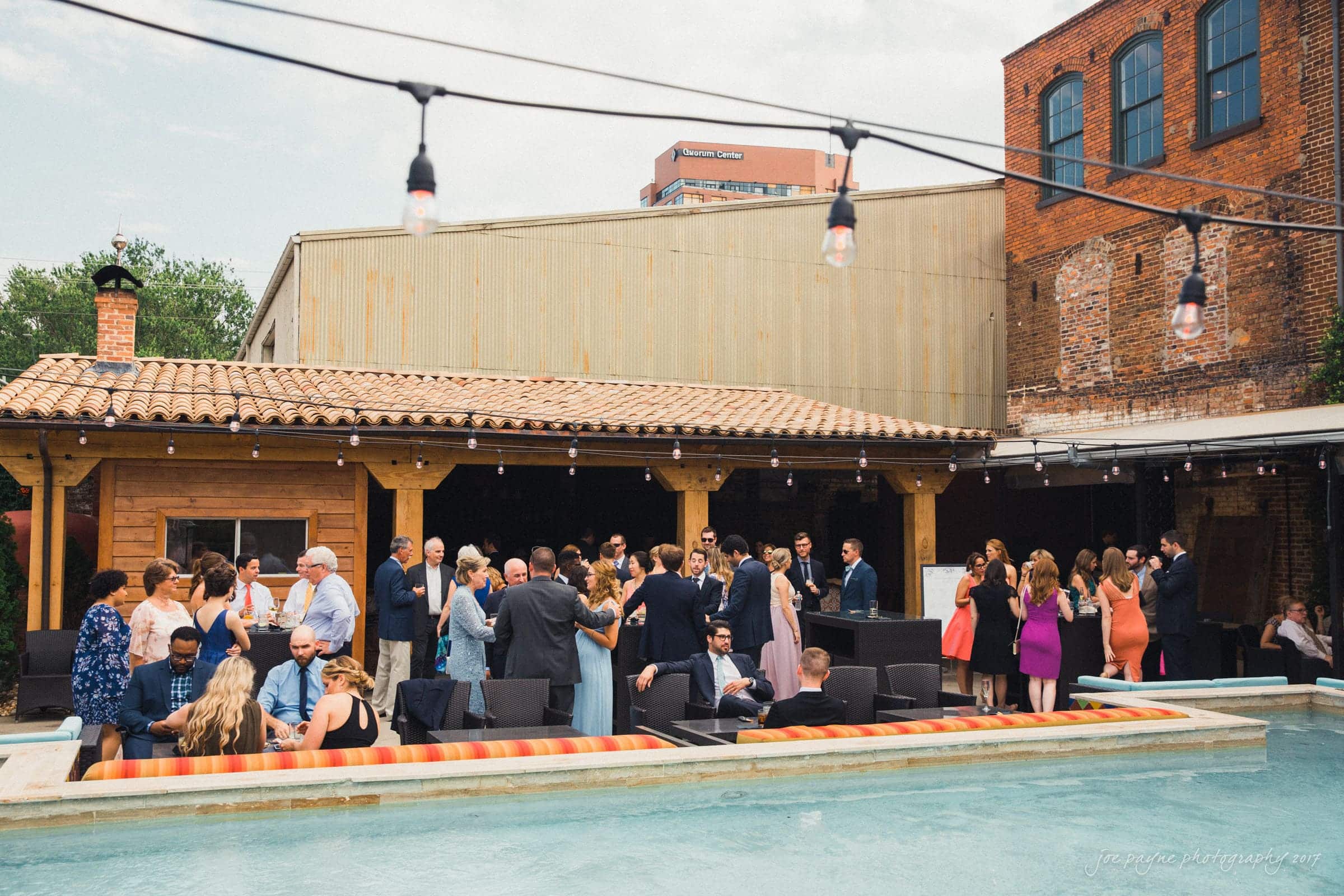 melrose knitting mill wedding - natalie & elliot A lively outdoor gathering with people socializing near a covered patio. String lights hang above, and a pool is in the foreground. Attendees are dressed in formal attire, mingling in a relaxed atmosphere beside a rustic brick and wood building.