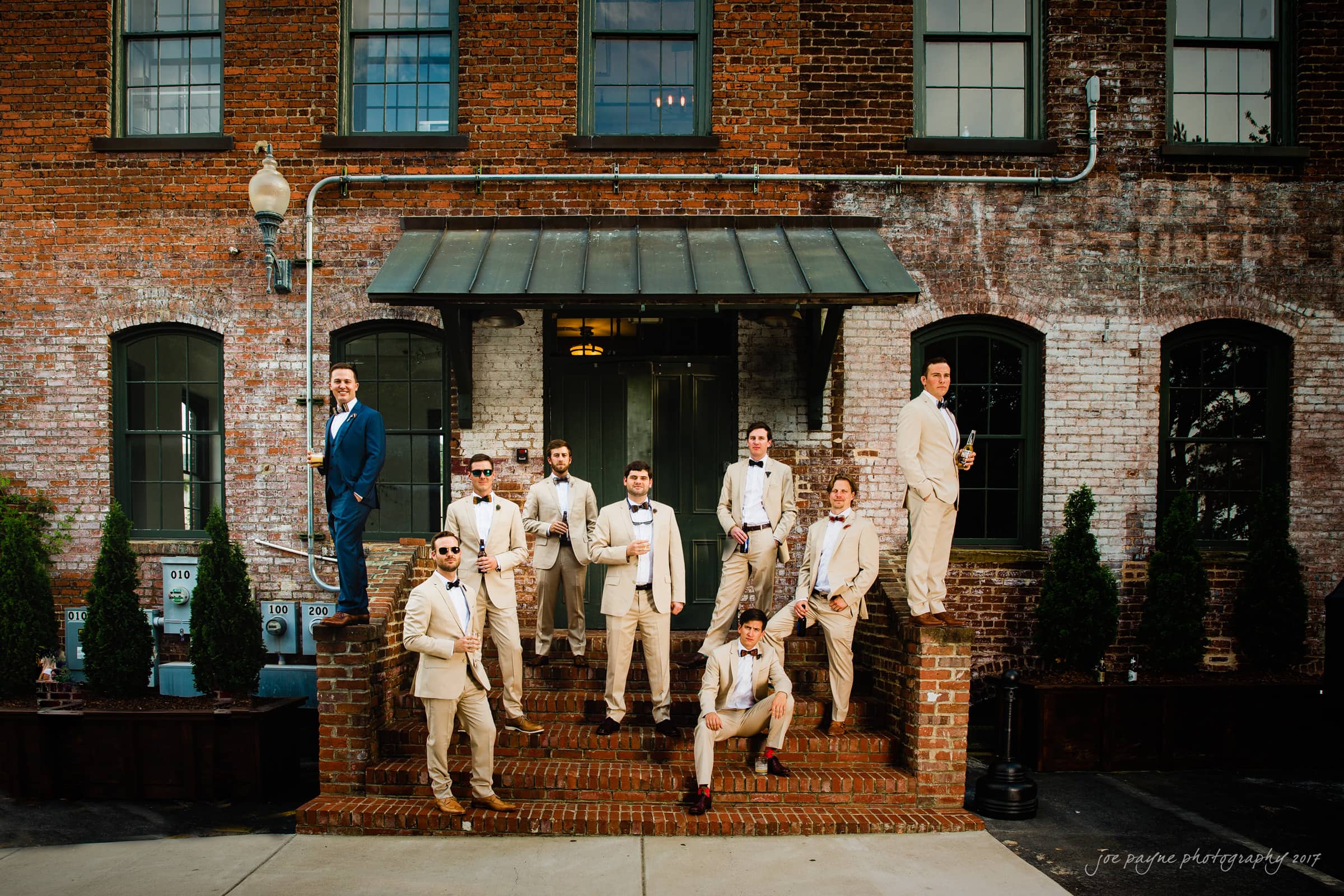 melrose knitting mill wedding - natalie & elliot A group of nine men in suits stands on the steps of a brick building. Some pose casually, while others lean against the wall. The setting is industrial with greenery visible in planters.