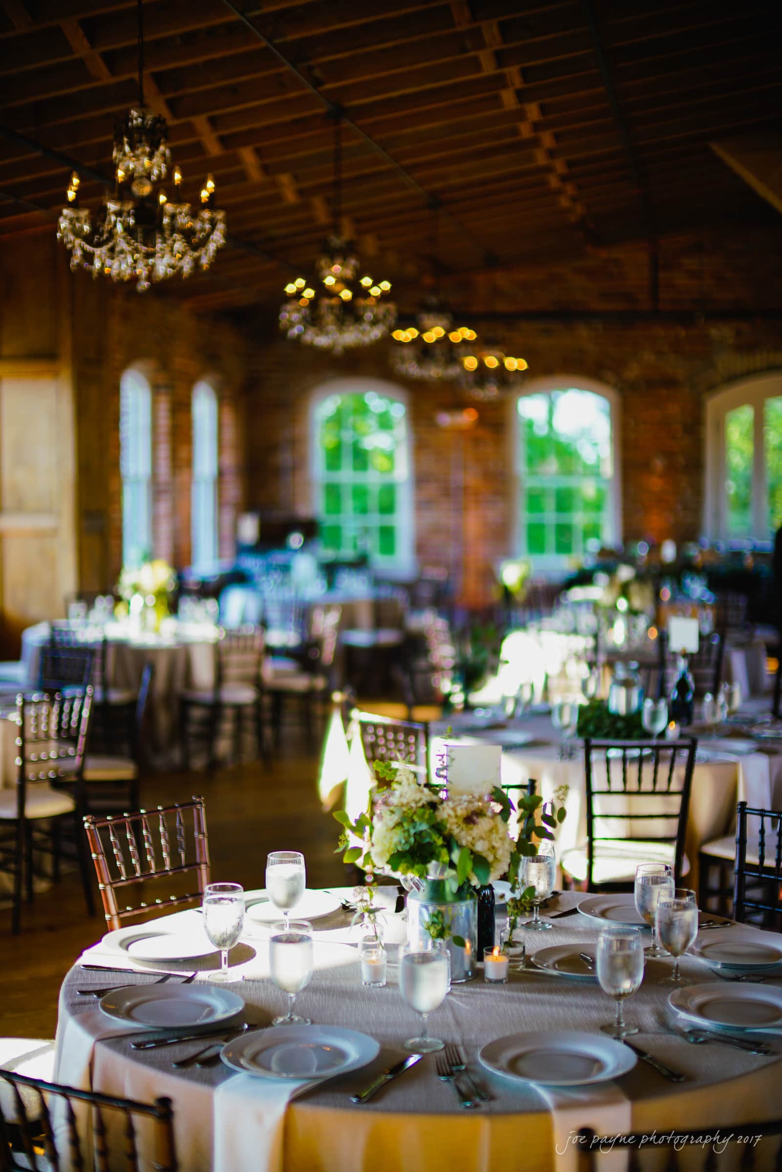 melrose knitting mill wedding - natalie & elliot A warmly lit dining room set up for an event with several round tables covered in white tablecloths, each adorned with place settings, glasses, and floral centerpieces. Elegant chandeliers hang from the wooden ceiling, and sunlight streams through large windows.