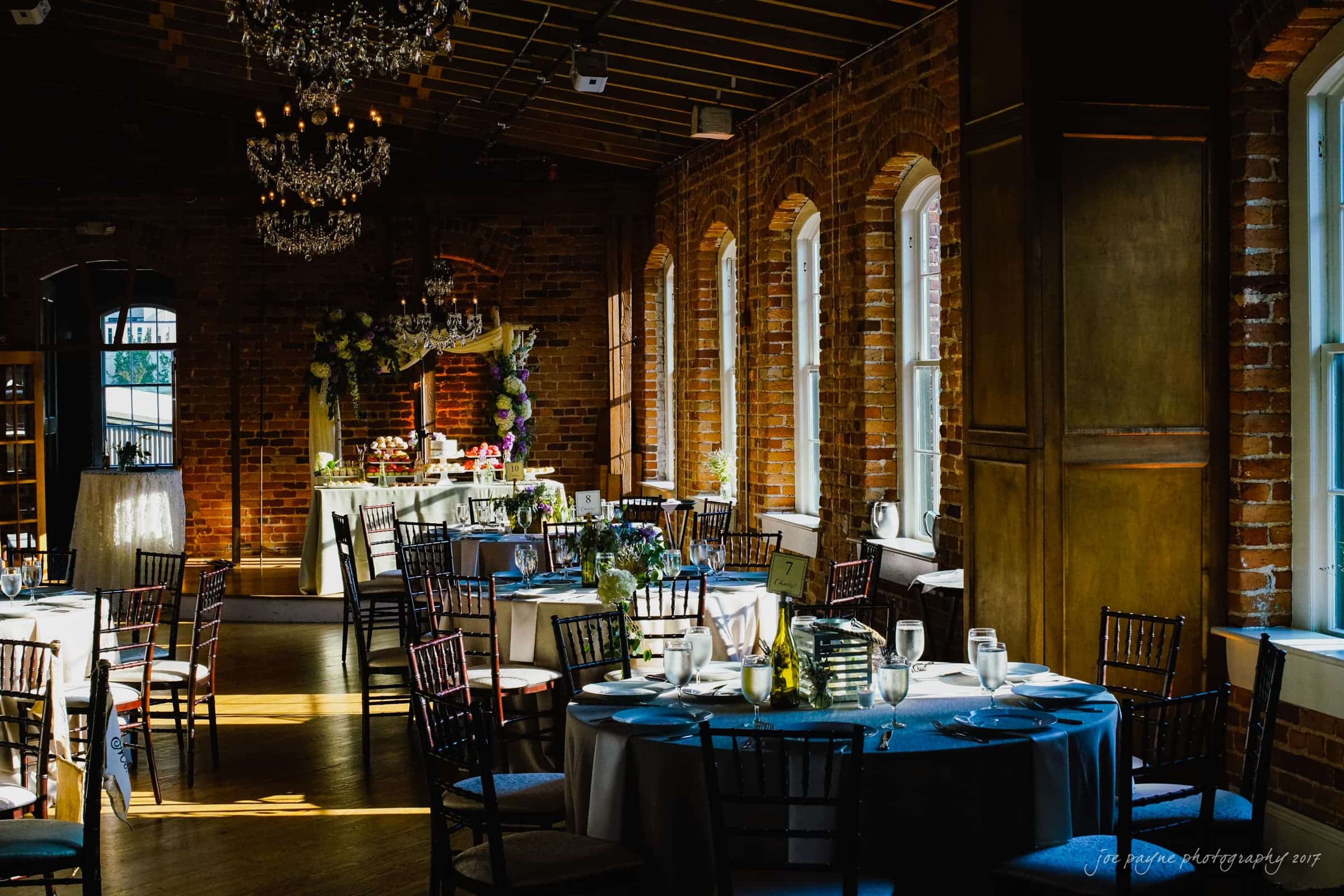 melrose knitting mill wedding - natalie & elliot A beautifully set indoor event space features round tables with white tablecloths and blue napkins. Sunlight streams through large arched windows, illuminating the brick walls and elegant chandeliers. Floral arrangements add a touch of color.
