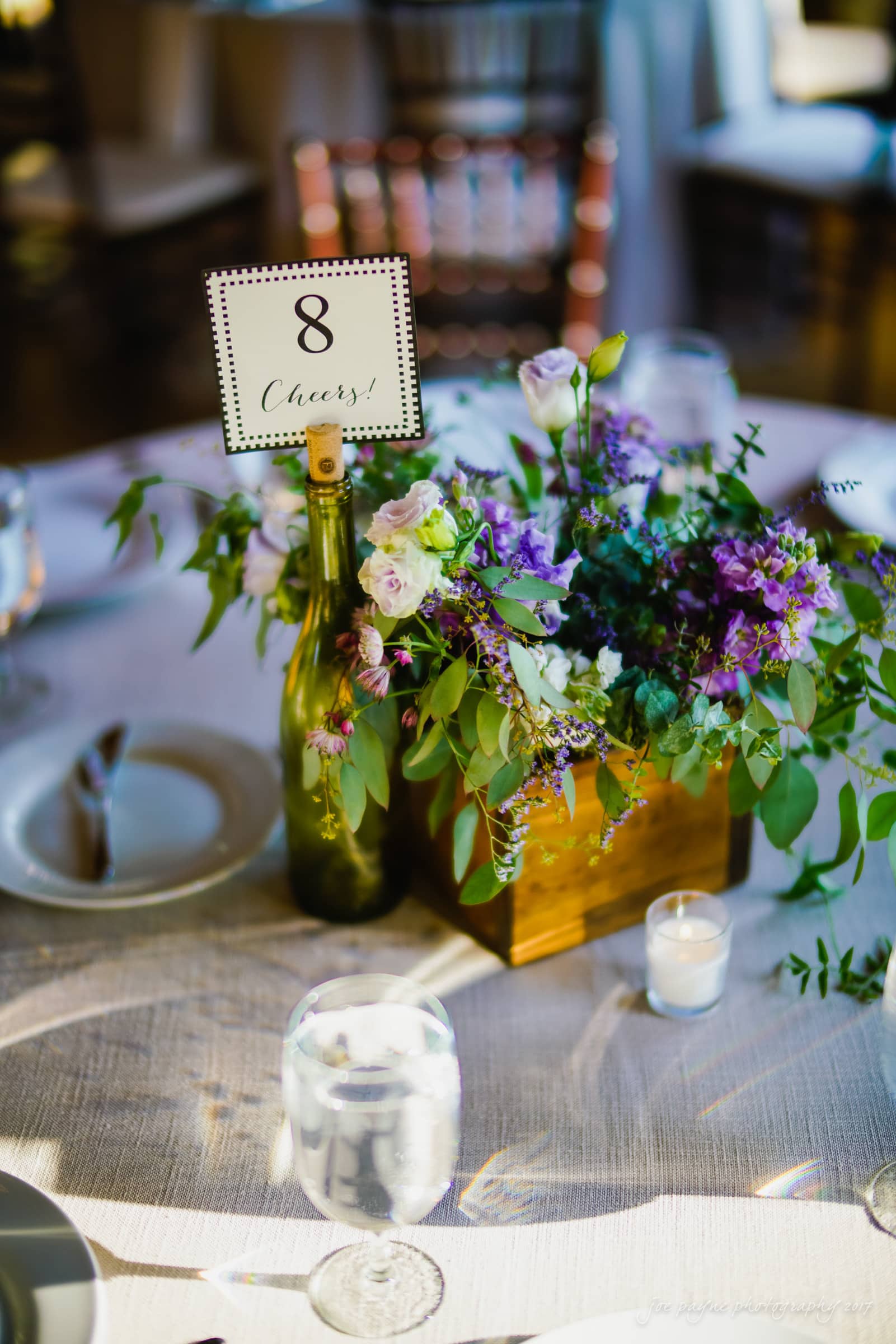 melrose knitting mill wedding - natalie & elliot A table setting with a centerpiece featuring a purple and white floral arrangement in a wooden box and a bottle holding a Table 8 Cheers! card. A small candle and glass of water are placed nearby on the tablecloth.