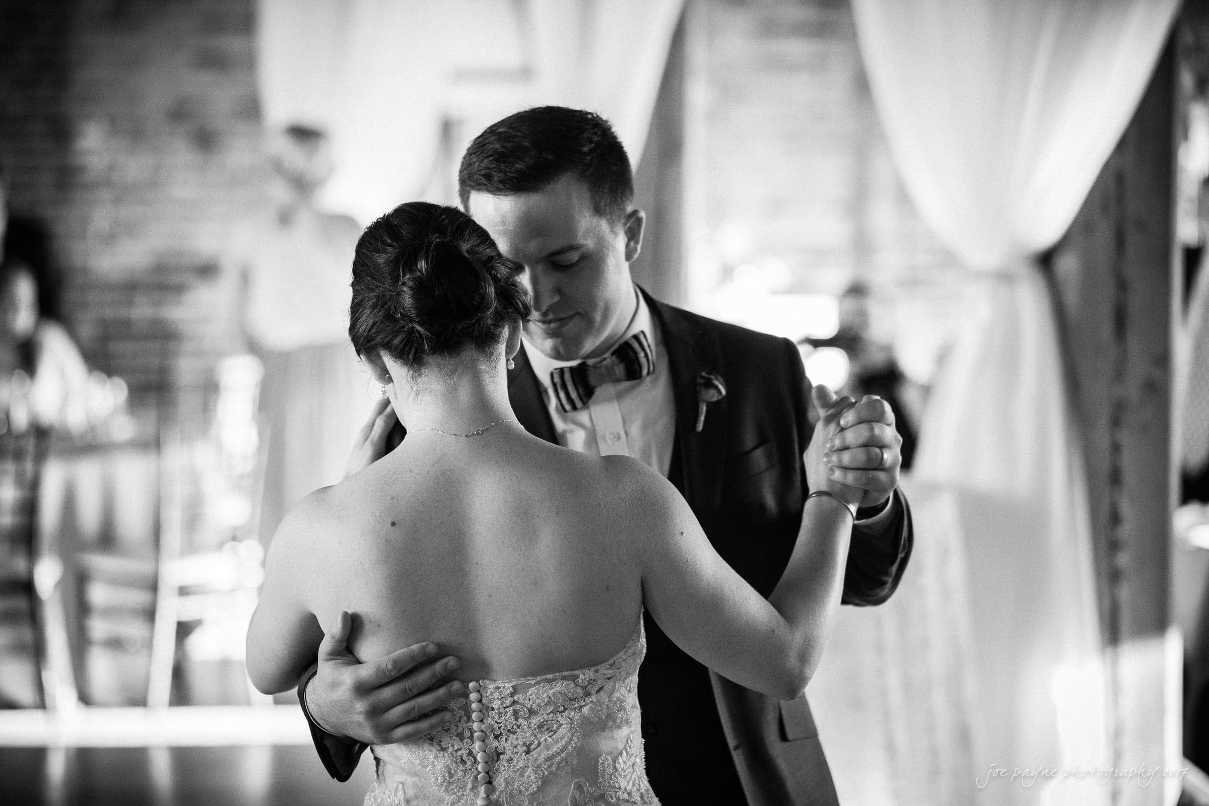 melrose knitting mill wedding - natalie & elliot A black and white photo of a couple dancing closely at an event. The man, wearing a suit and bow tie, gently holds the woman, who is in a strapless dress. Soft curtains and brick walls are visible in the background, creating a romantic atmosphere.