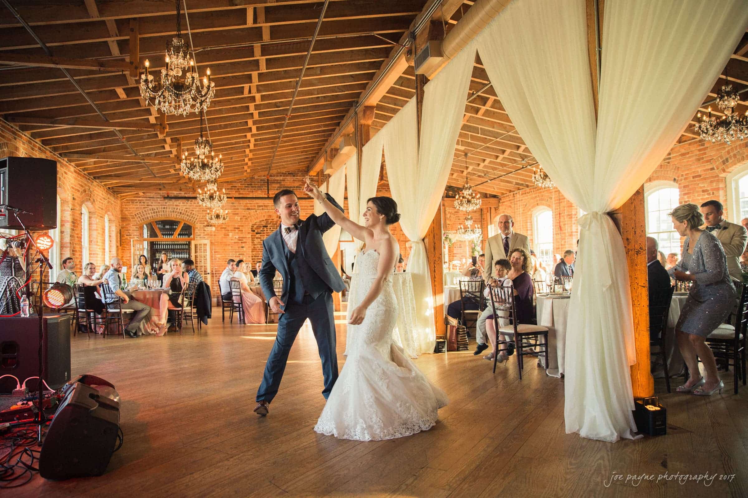 melrose knitting mill wedding - natalie & elliot A couple in wedding attire dances in a brick-walled venue with guests seated at tables. Chandeliers hang from the ceiling, and draped fabric decorates the space. Natural light fills the room.