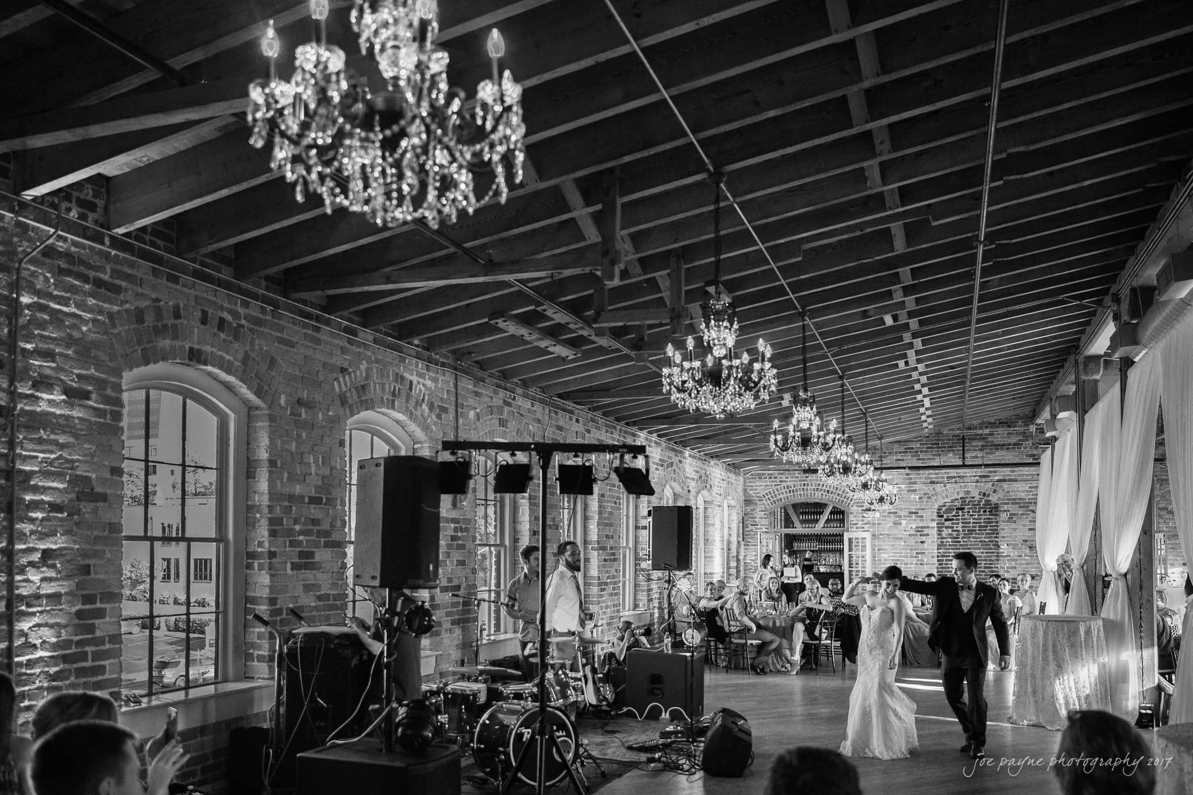 melrose knitting mill wedding - natalie & elliot A black and white photo of a couple dancing joyfully in a large, rustic hall with exposed brick walls and wooden beams. Chandeliers hang from the ceiling, and guests are seated at tables, watching the dance. A live band setup is visible.