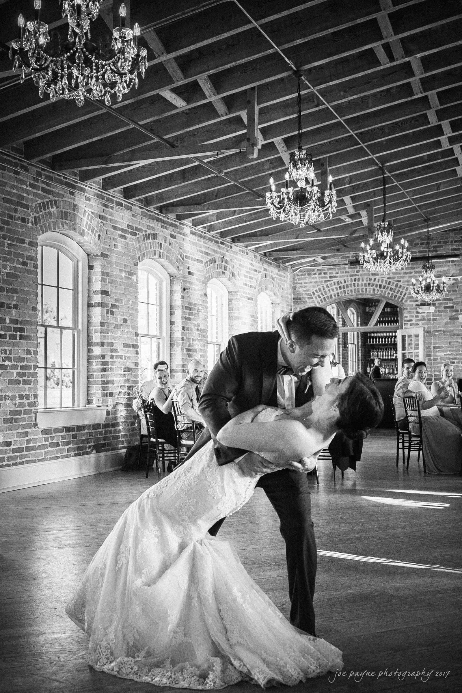 melrose knitting mill wedding - natalie & elliot A couple in formal attire shares a romantic dance and kiss in a rustic venue with exposed brick walls and chandeliers. They are surrounded by seated guests who observe the scene, creating a warm and celebratory atmosphere.