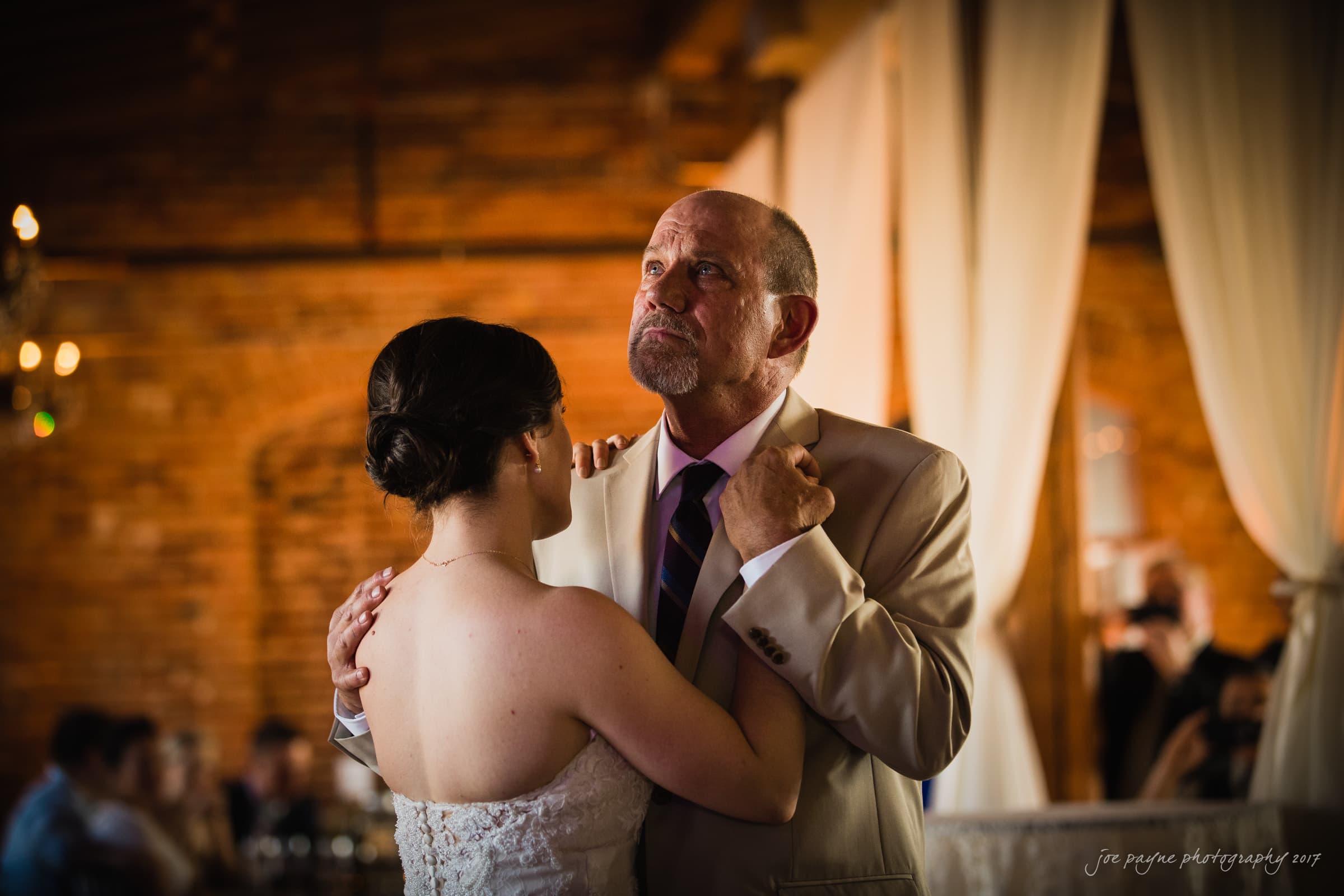 melrose knitting mill wedding - natalie & elliot A bride and an older man are dancing together in a warmly lit, rustic setting. The bride wears a white dress, and the man is in a light-colored suit. Both gaze upwards, creating a serene and intimate atmosphere.