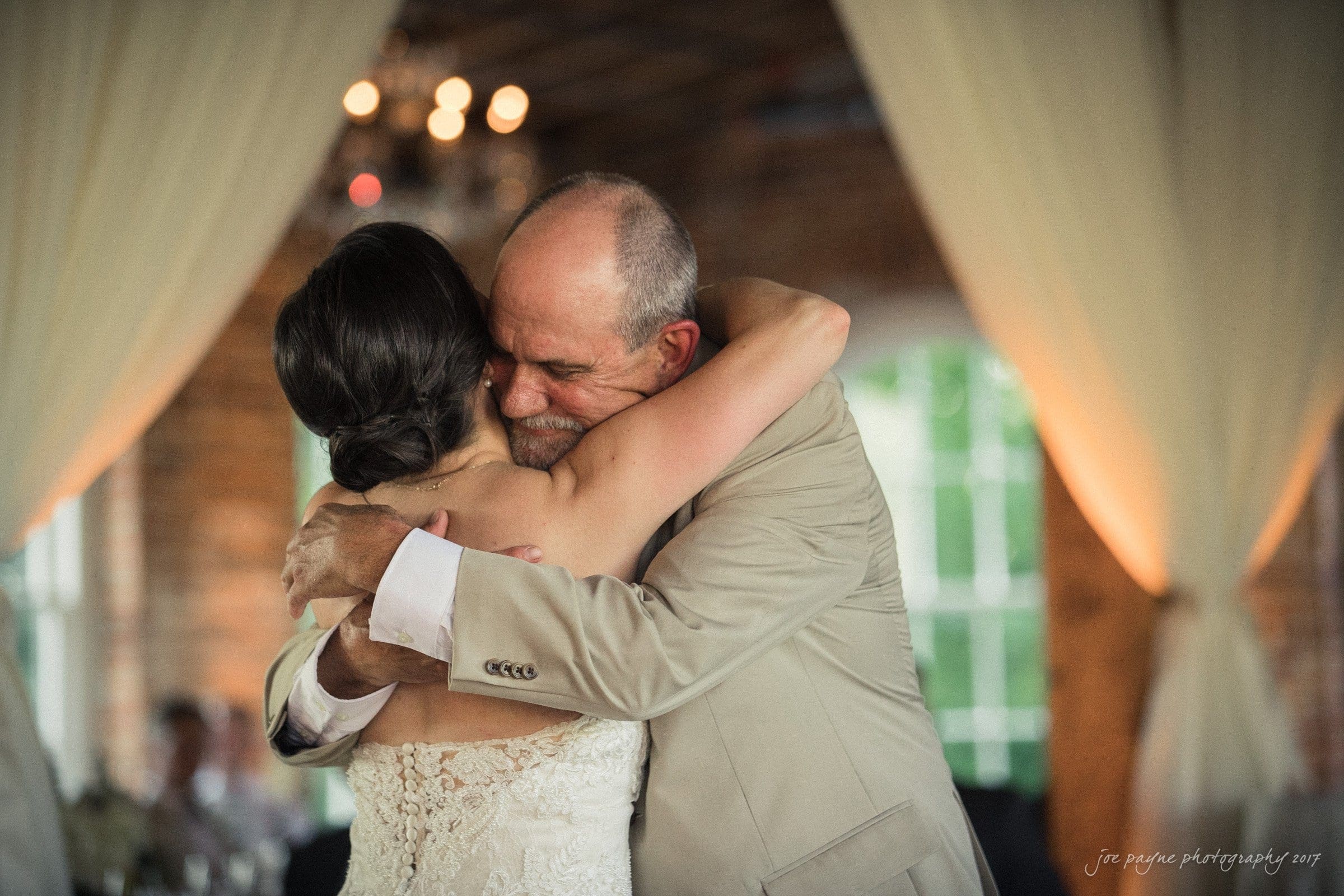 melrose knitting mill wedding - natalie & elliot A man and a woman embrace warmly at what appears to be a wedding, surrounded by light curtains and a softly lit room. The mans eyes are closed in a tender moment, and the womans back is toward the camera.