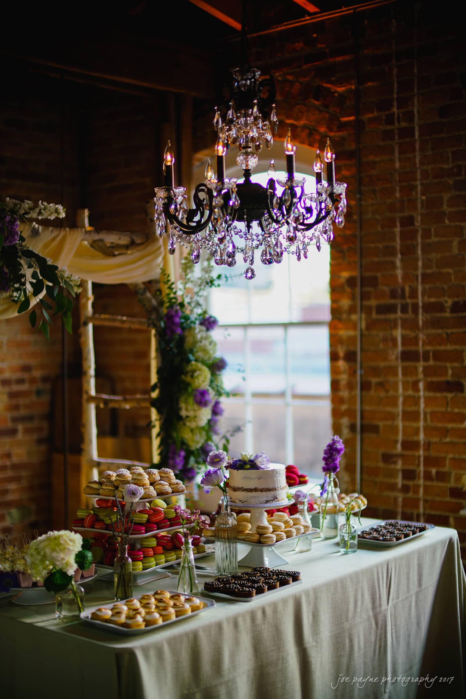 melrose knitting mill wedding - natalie & elliot A wedding dessert table decorated with purple and white flowers. It features a tiered cake, stacks of macarons, and trays of chocolates. A chandelier above casts a warm glow in a rustic brick setting.