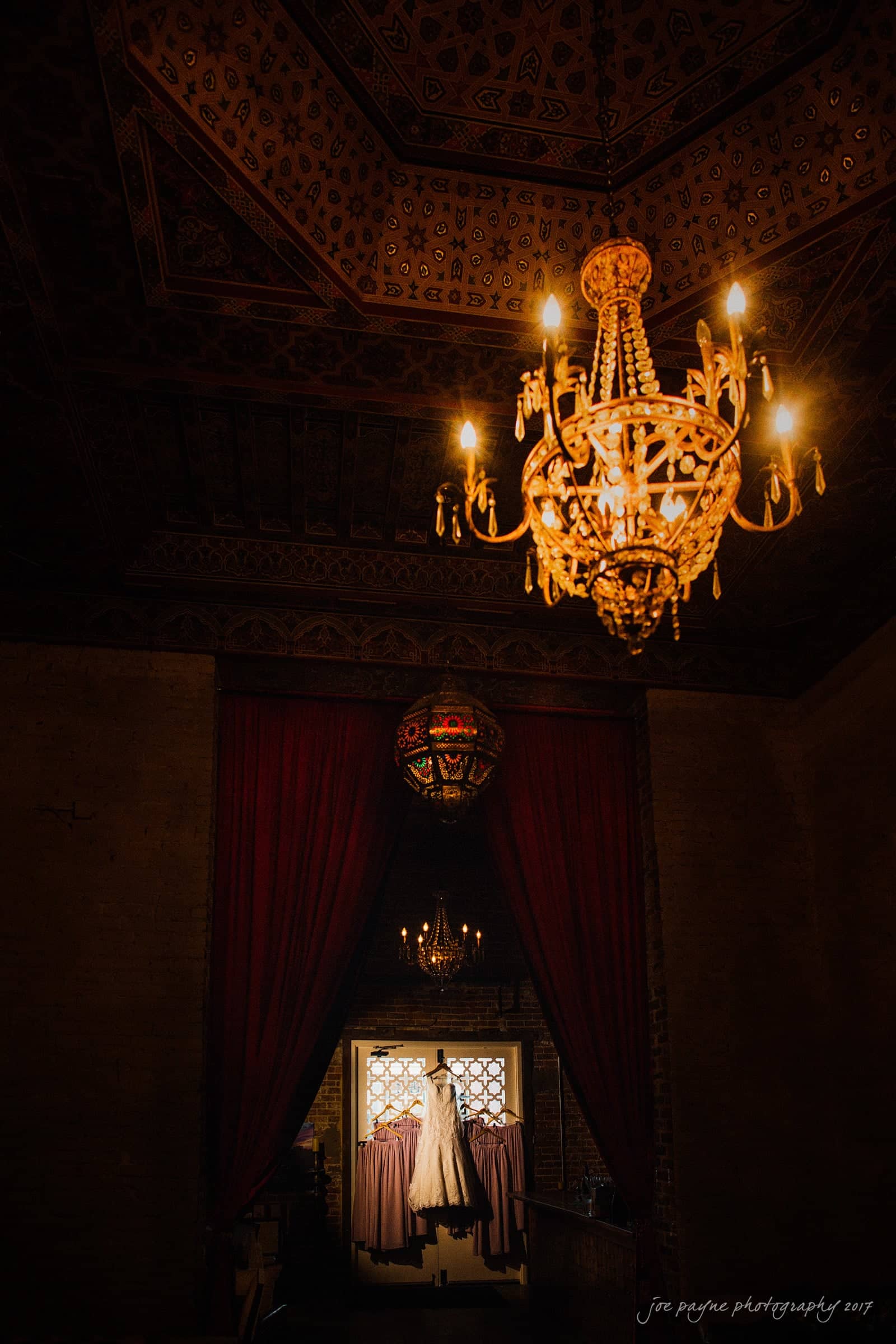 melrose knitting mill wedding - natalie & elliot A vintage chandelier illuminates a dimly lit room with ornate ceiling patterns. Below, a wedding dress is displayed on an elevated platform, framed by rich red curtains and a decorative window in the background.