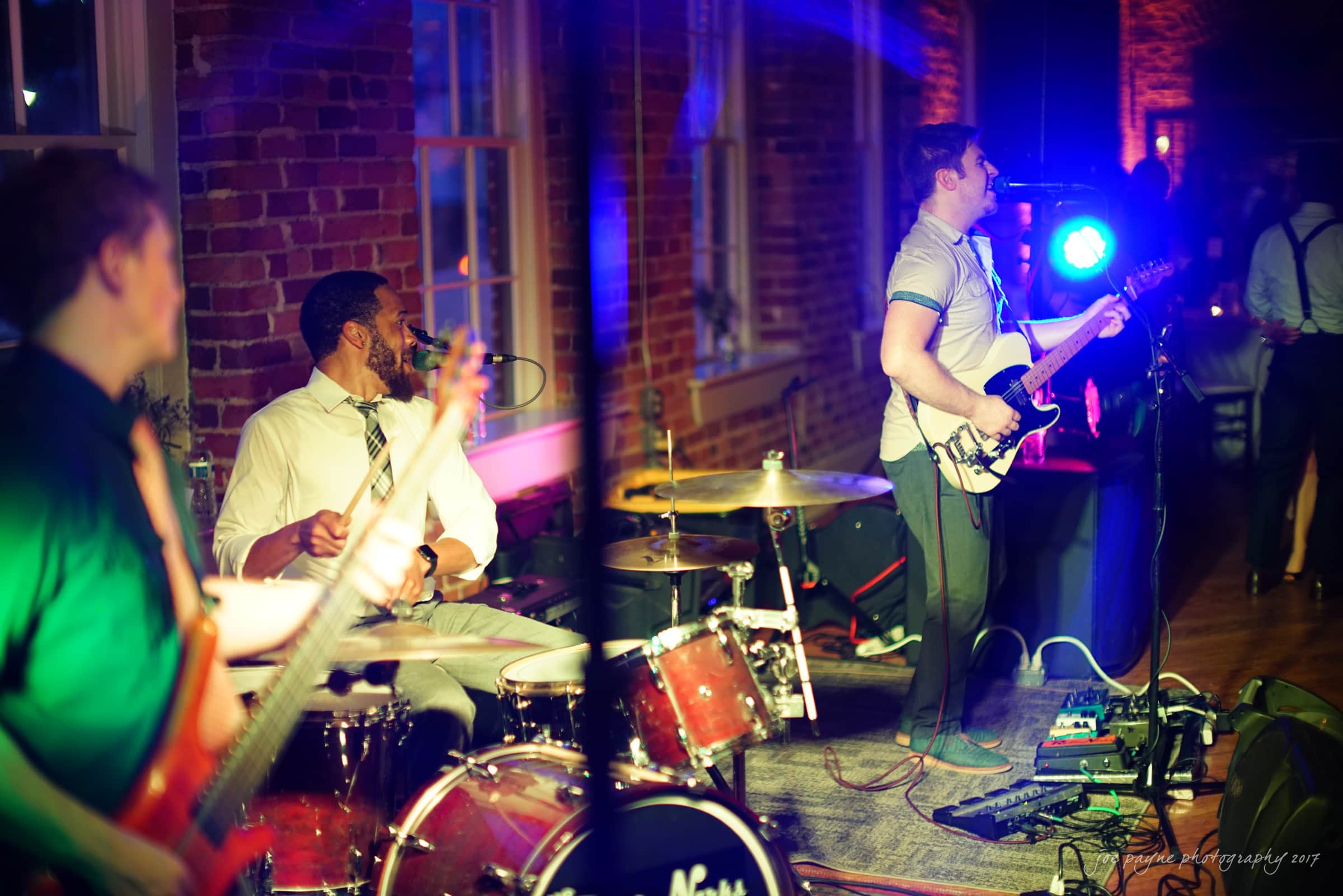 melrose knitting mill wedding - natalie & elliot A live band performs on stage in a dimly lit venue. The guitarist sings into a microphone while playing, and the drummer behind him is also visible. Colorful stage lights illuminate the scene, reflecting off the brick walls.