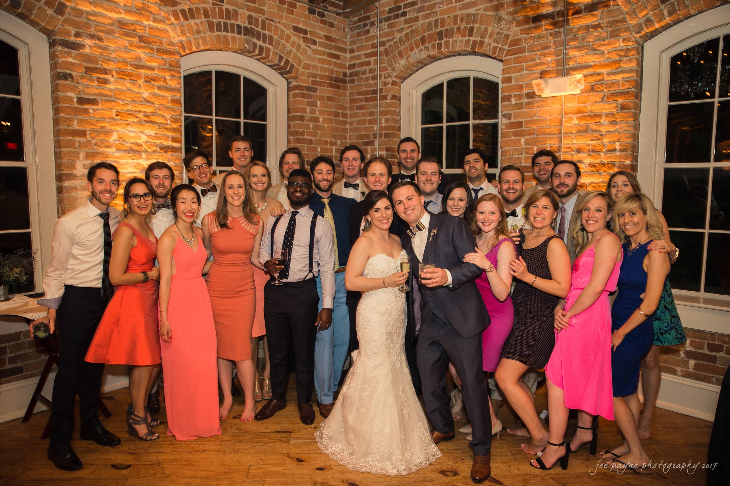 melrose knitting mill wedding - natalie & elliot A large group of people, dressed in formal attire, are gathered indoors for a celebration. The couple in the center is wearing wedding attire, with the bride in a white gown and the groom in a suit. The room features exposed brick walls and wooden floors.