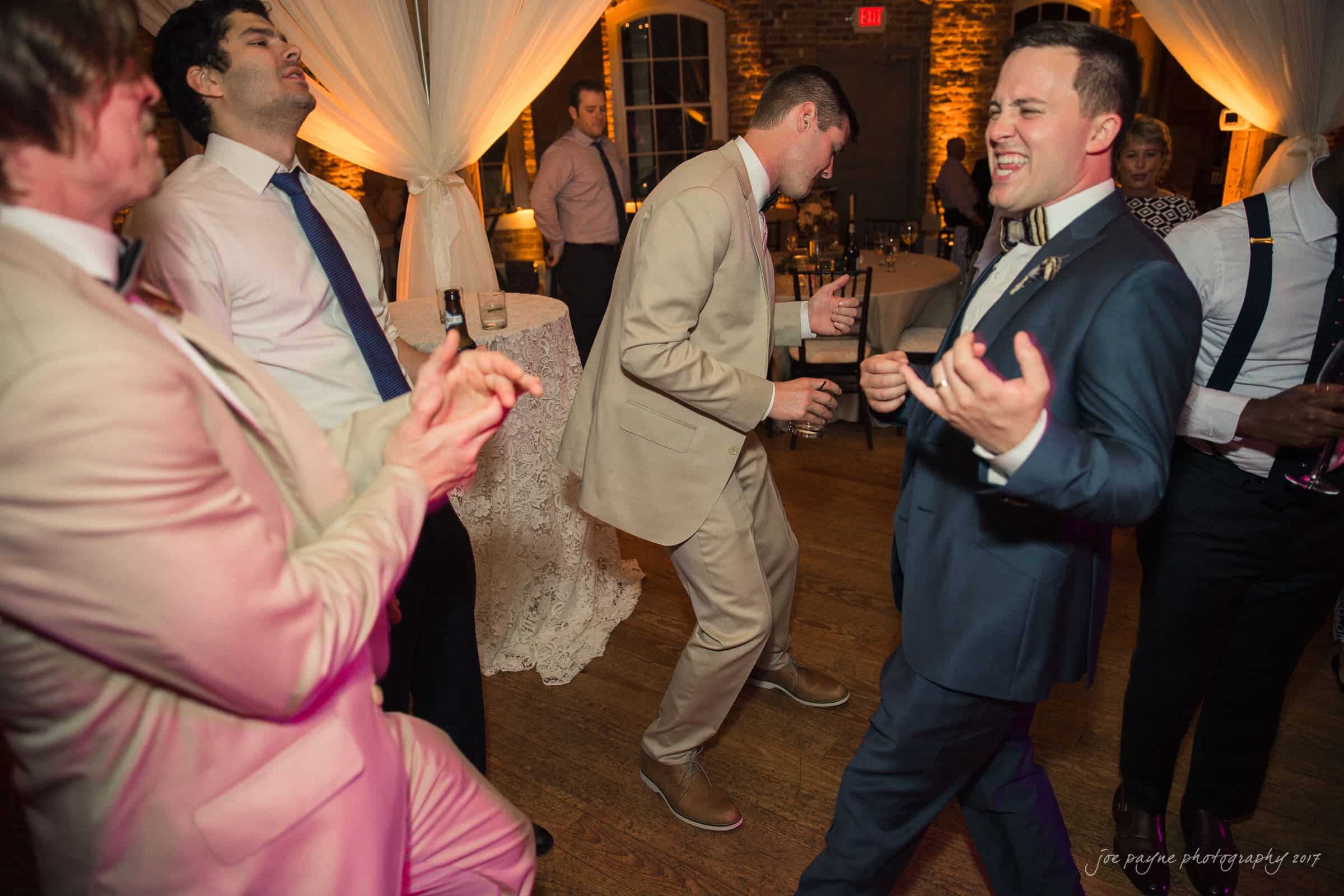 melrose knitting mill wedding - natalie & elliot A group of men in suits dance enthusiastically in a warmly lit, elegant venue. They appear joyful, enjoying the moment on a wooden floor with draped curtains and soft lighting in the background.