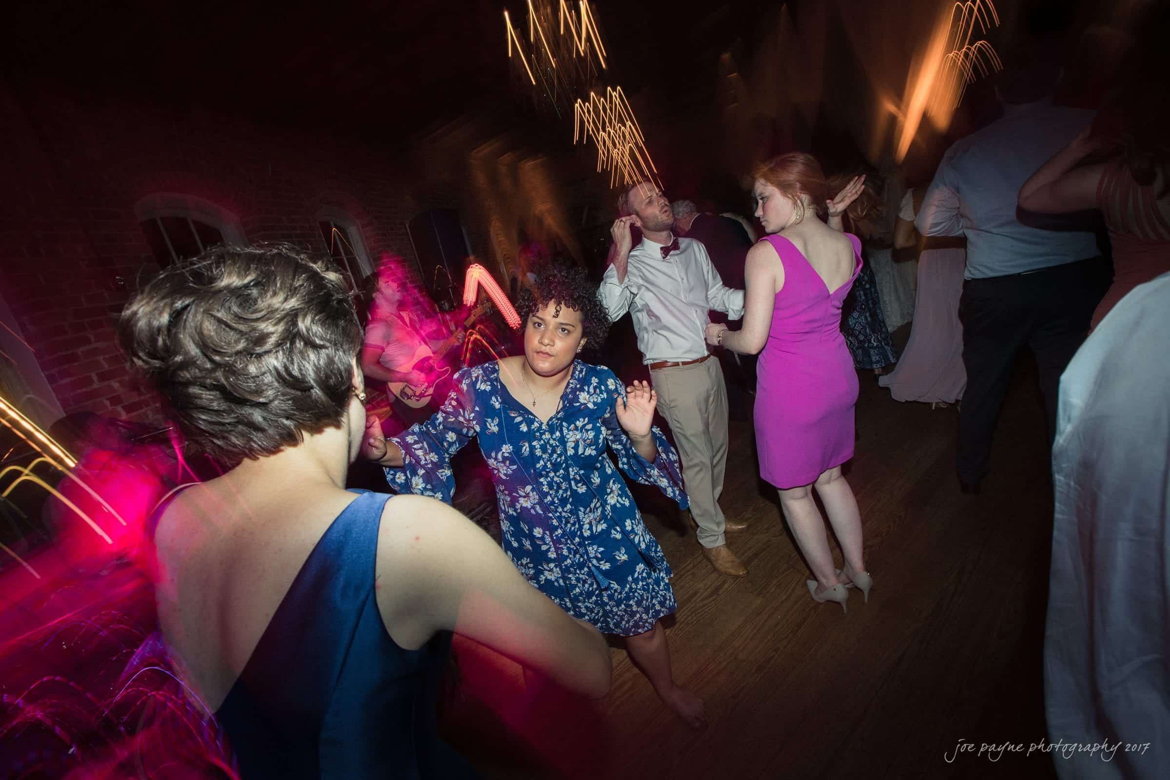 melrose knitting mill wedding - natalie & elliot People are dancing energetically in a dimly lit room with blurred light streaks. A woman in a blue floral dress is in focus, surrounded by others in formal attire. The atmosphere is lively and festive.