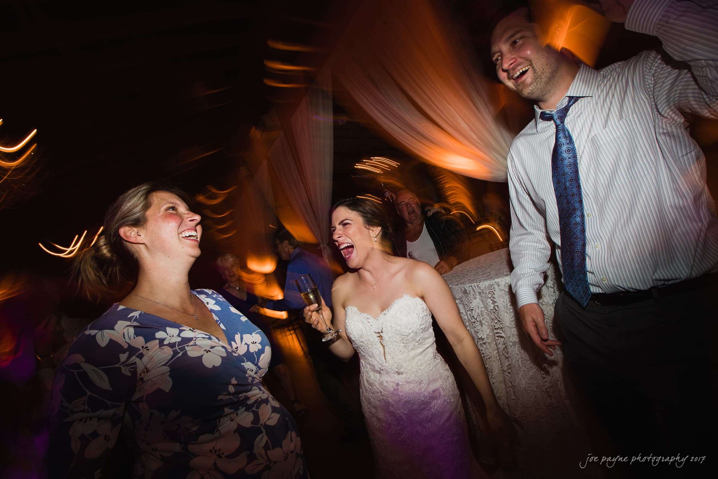 melrose knitting mill wedding - natalie & elliot A joyful wedding scene with a bride in a white dress laughing and dancing with two guests. One guest wears a floral dress, and the other a shirt and tie. The room is warmly lit, creating a vibrant atmosphere.