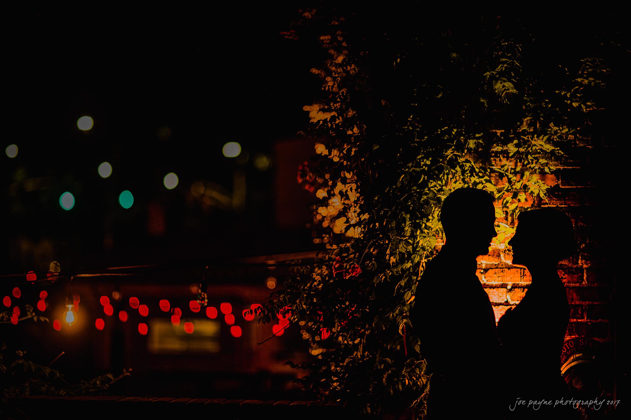 melrose knitting mill wedding - natalie & elliot Silhouetted couple facing each other in front of a brick wall adorned with foliage and warm yellow lighting. Red string lights are visible in the background, creating a romantic nighttime ambiance.