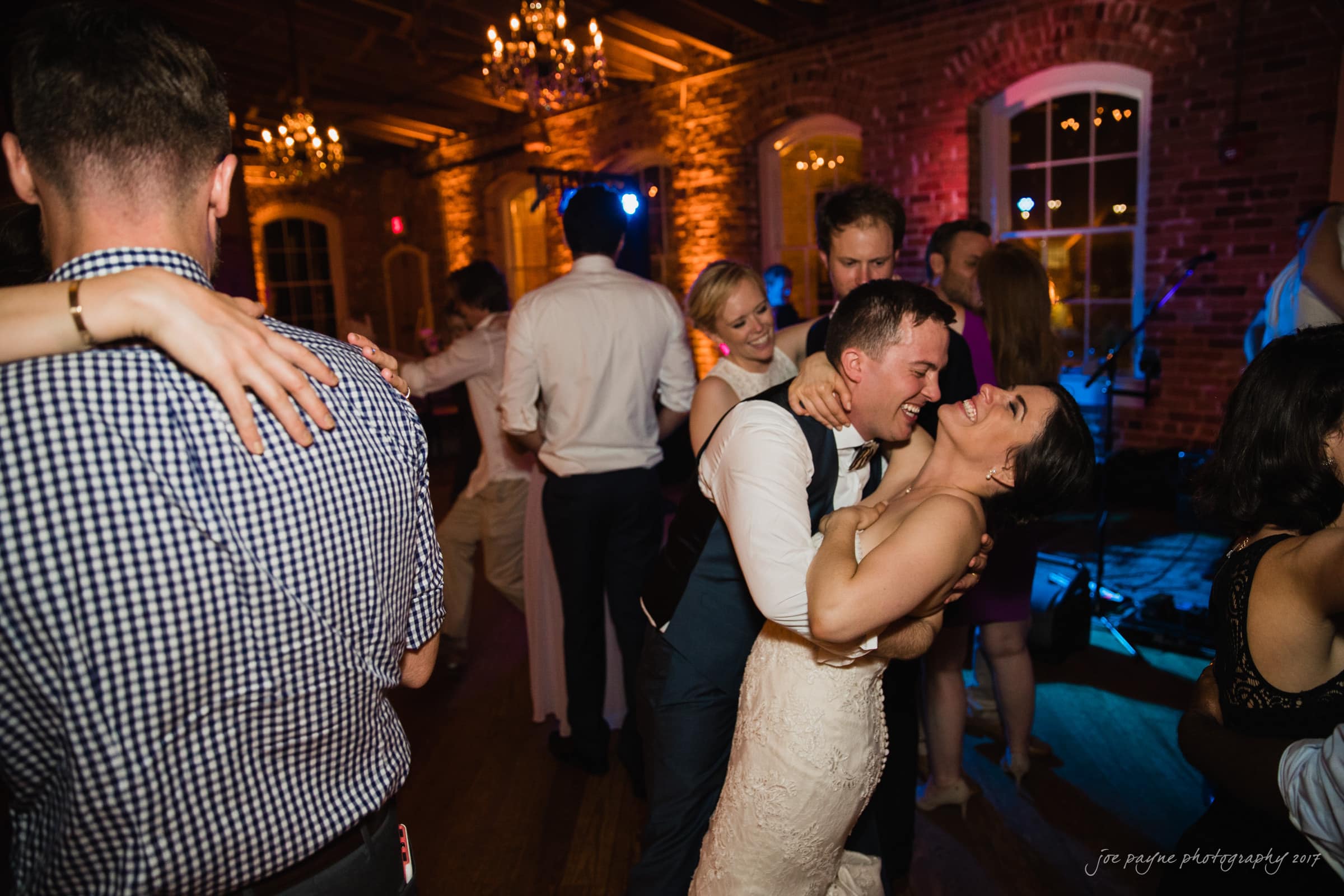 melrose knitting mill wedding - natalie & elliot A joyful couple dances closely at a lively event in a dimly lit venue with exposed brick walls and chandeliers. Other guests dance and socialize around them.