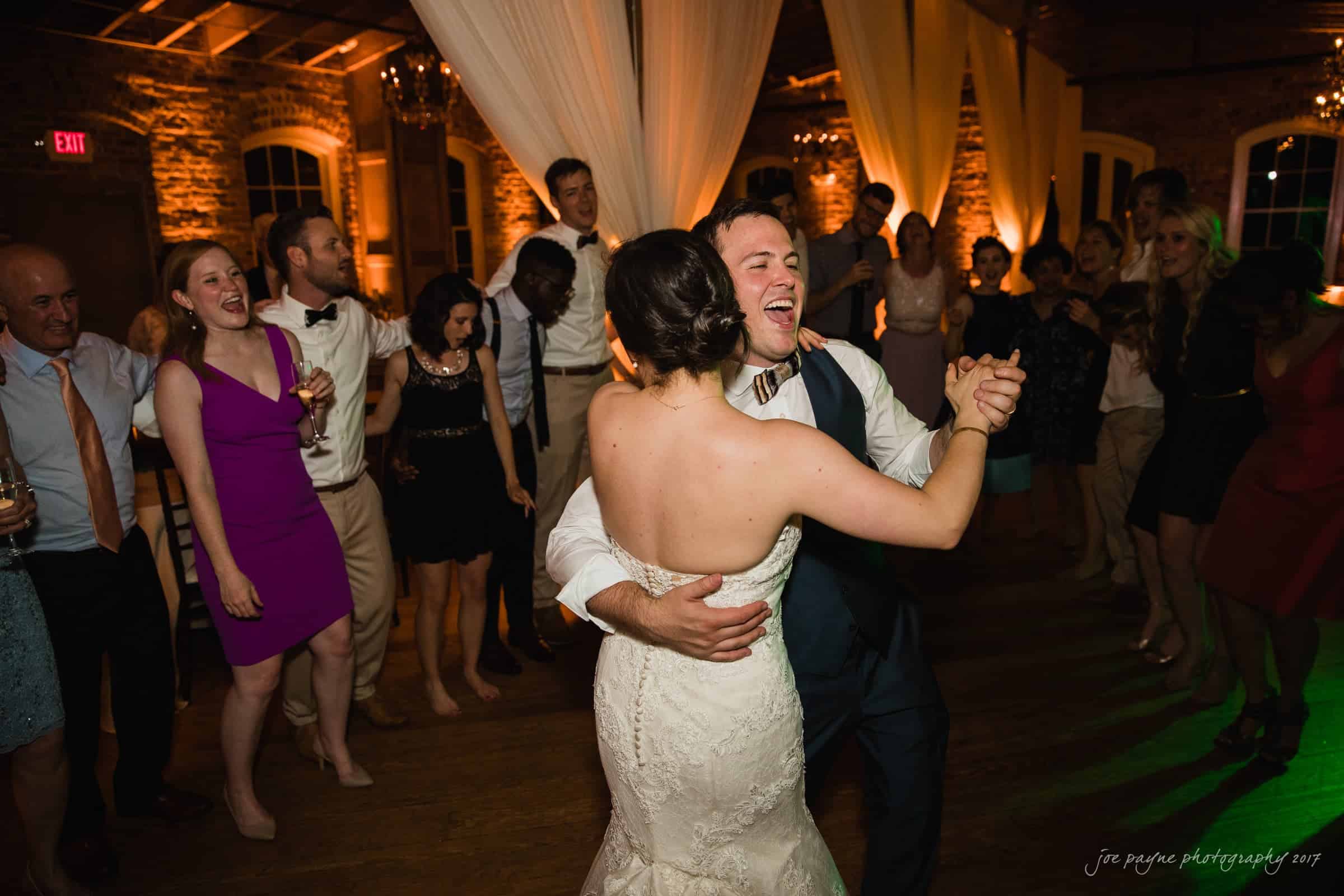 melrose knitting mill wedding - natalie & elliot A bride and groom dance joyfully together at a wedding reception, surrounded by smiling guests dressed in formal attire. The warmly lit venue has brick walls and draped white curtains, creating a festive atmosphere.