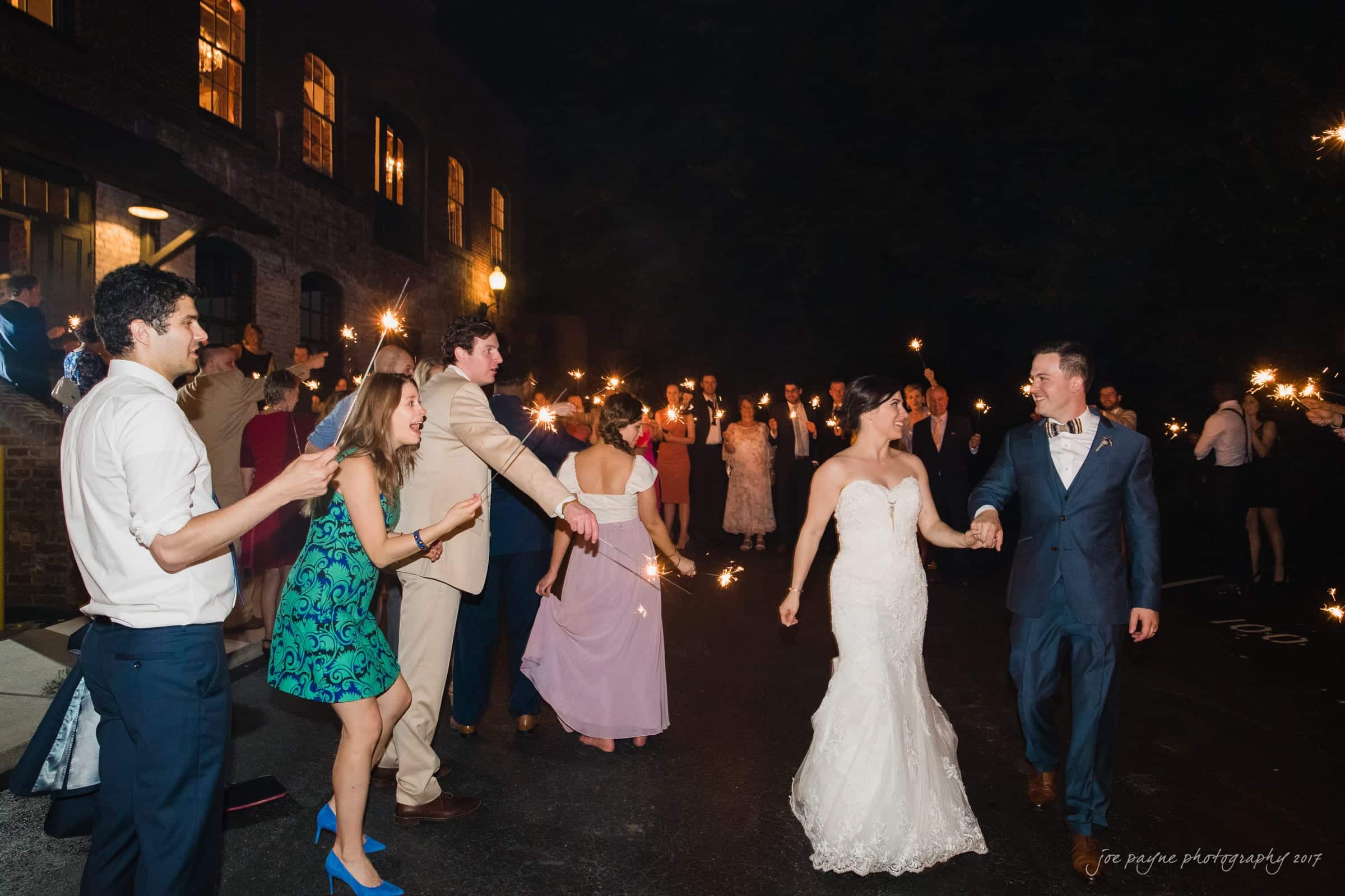 melrose knitting mill wedding - natalie & elliot Bride and groom walk hand in hand through a corridor of guests holding sparklers at night. The bride wears a white gown and the groom a dark suit. Guests cheer and celebrate as they pass by.