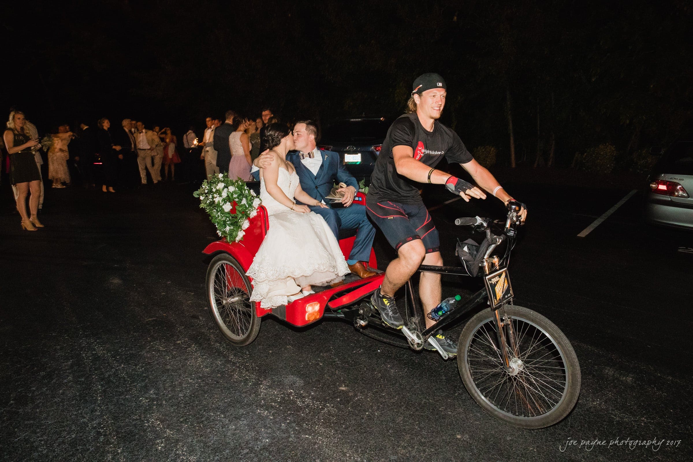 melrose knitting mill wedding - natalie & elliot A bride and groom share a joyful moment on a pedicab at night. The groom kisses the brides cheek, and shes holding a bouquet. A cyclist pedals them forward. Guests are seen in the background.