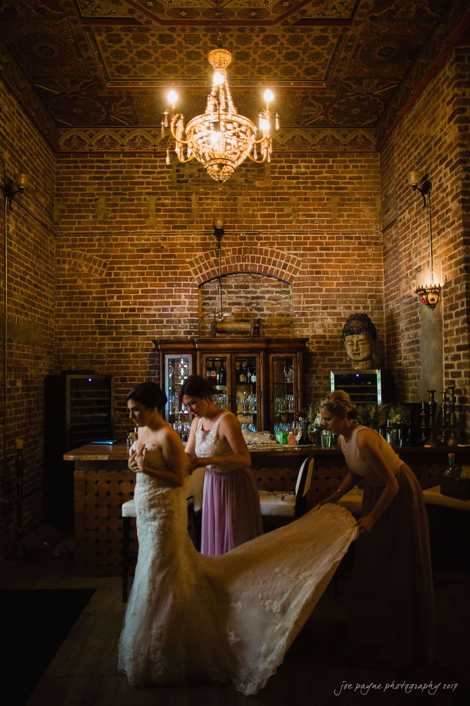 melrose knitting mill wedding - natalie & elliot A bride in a lace gown is assisted by two bridesmaids in a rustic room with exposed brick walls and a decorative ceiling. A chandelier hangs above, and a bar with a Buddha head sculpture is in the background.
