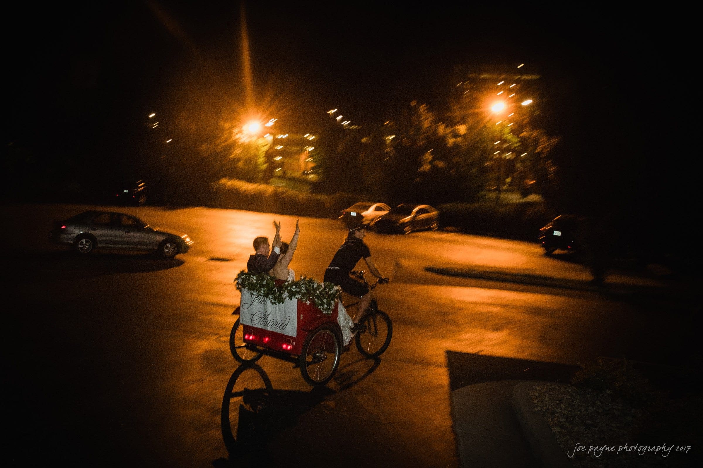 melrose knitting mill wedding - natalie & elliot A couple rides in a floral-decorated rickshaw at night, waving as they travel through a dimly lit parking lot surrounded by parked cars and illuminated by streetlights.