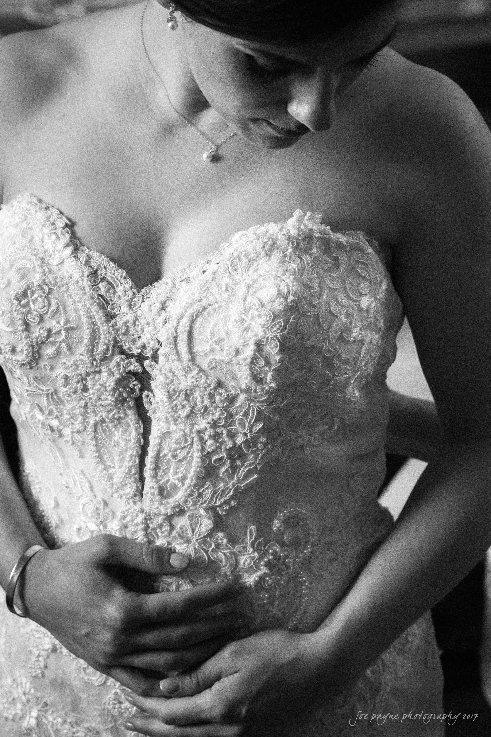 melrose knitting mill wedding - natalie & elliot Black and white photo of a bride in a detailed lace wedding dress. She stands with her head slightly bowed, as someone embraces her from behind, showcasing the intricate design of her gown.
