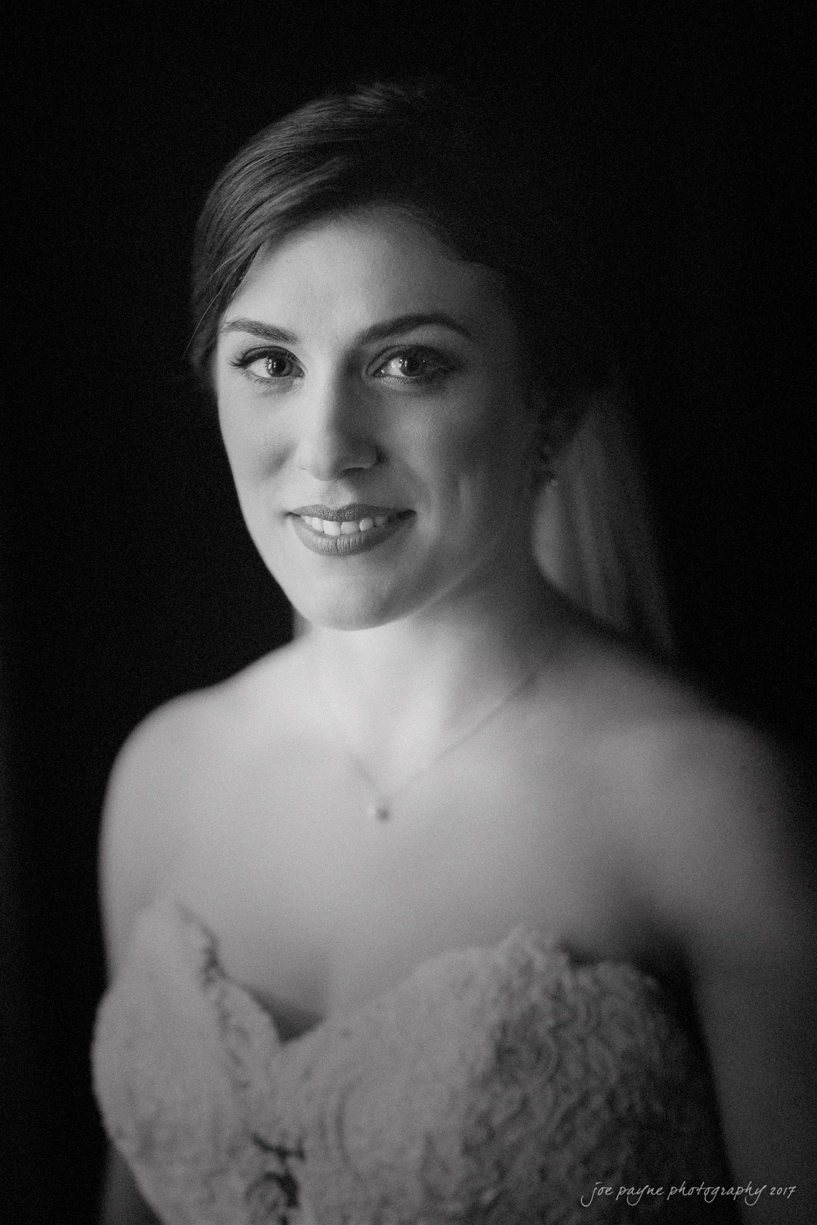 melrose knitting mill wedding - natalie & elliot Black and white portrait of a woman wearing a strapless lace wedding gown. She is smiling softly, looking into the camera, with a dark background that highlights her face.