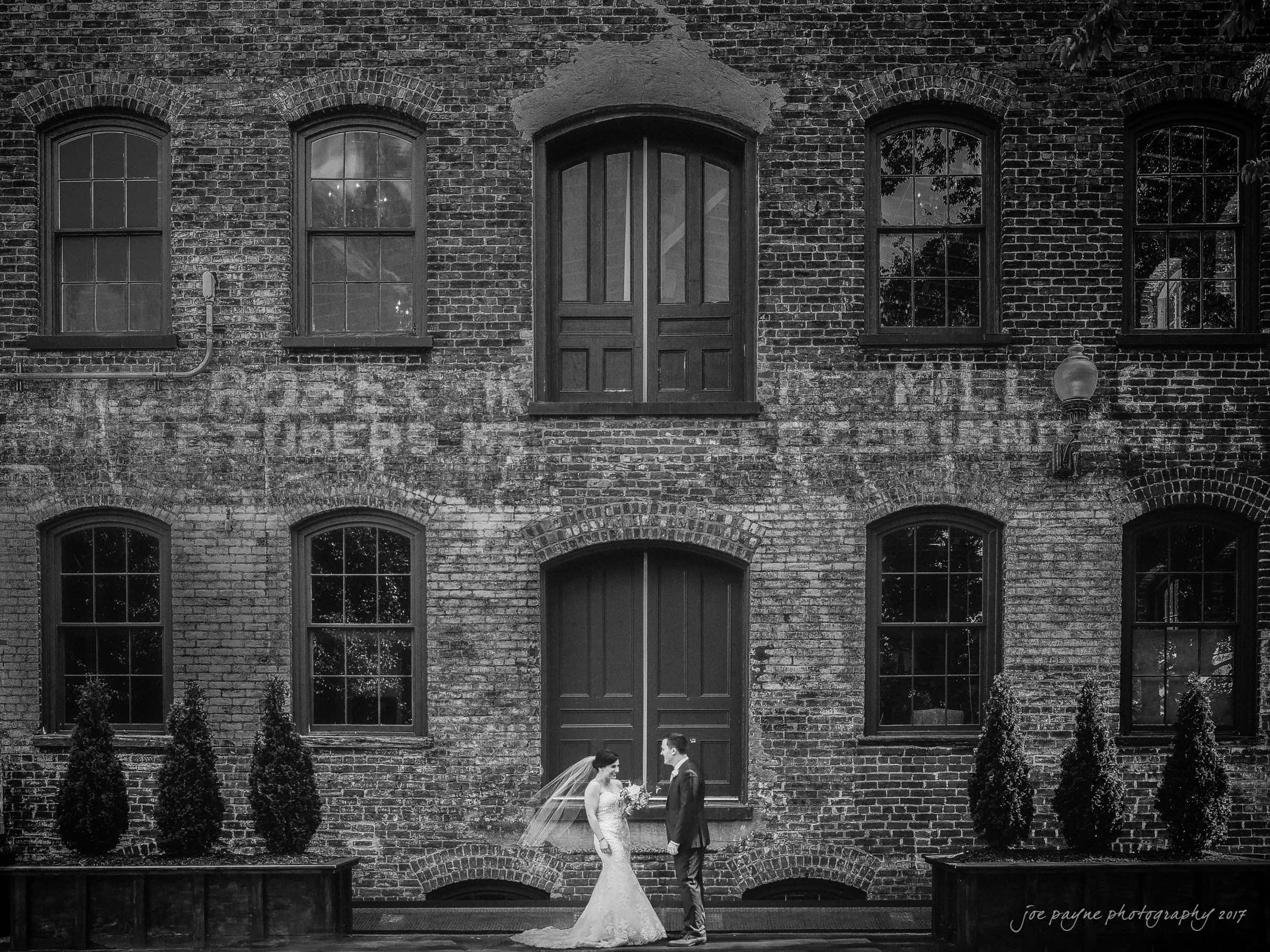 melrose knitting mill wedding - natalie & elliot A bride and groom stand facing each other in front of a large, historic brick building with tall windows. The bride holds a veil, and small trees line the foreground, creating a romantic atmosphere.