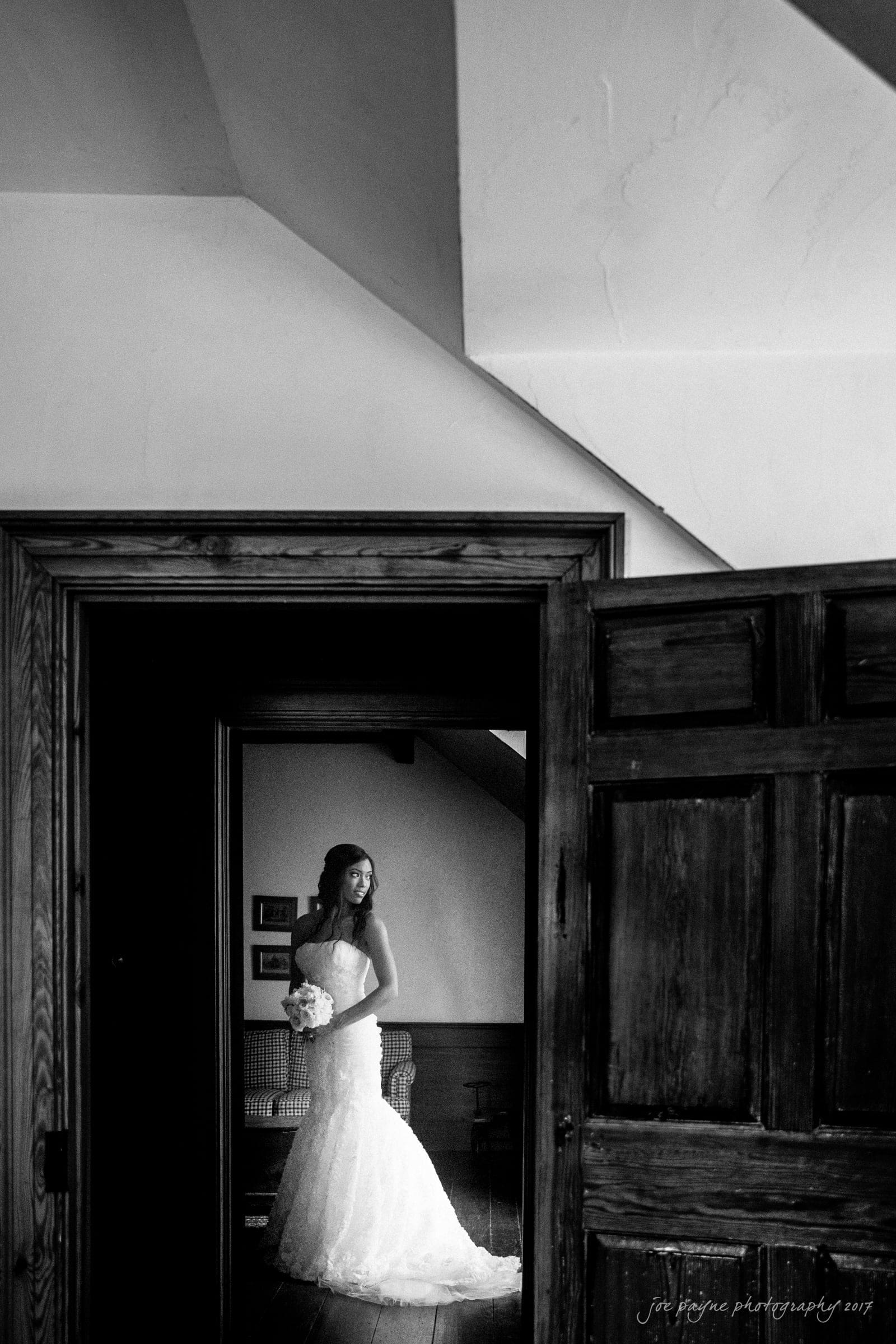 A bride in a strapless lace gown holds a bouquet, standing gracefully in a dimly lit room. She gazes out of a wooden doorway, surrounded by rustic decor. The atmosphere is serene and contemplative.