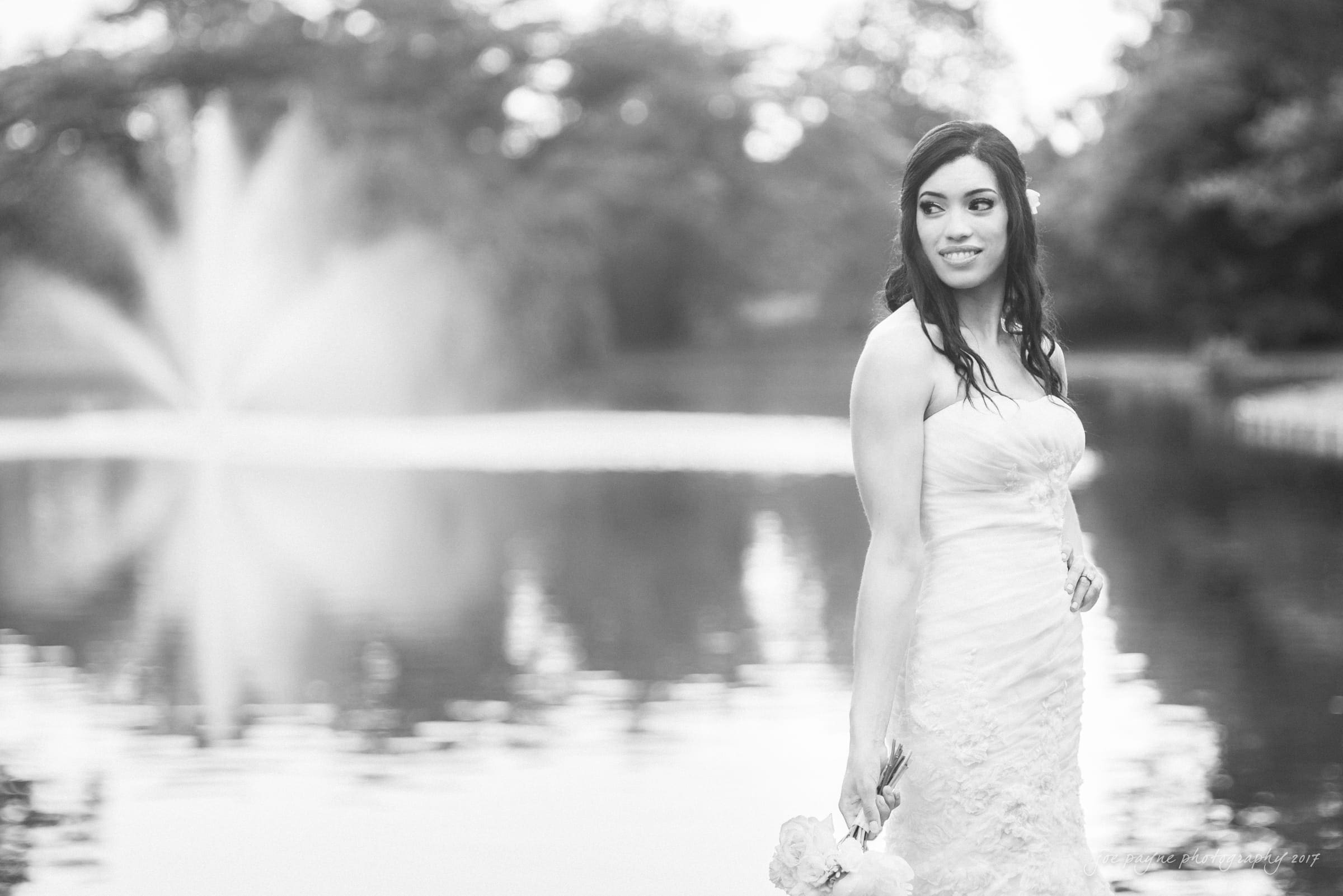 A bride in a strapless wedding dress stands by a lake, holding a bouquet. The background features a fountain and blurred greenery, creating a serene and elegant scene. The image is in black and white.