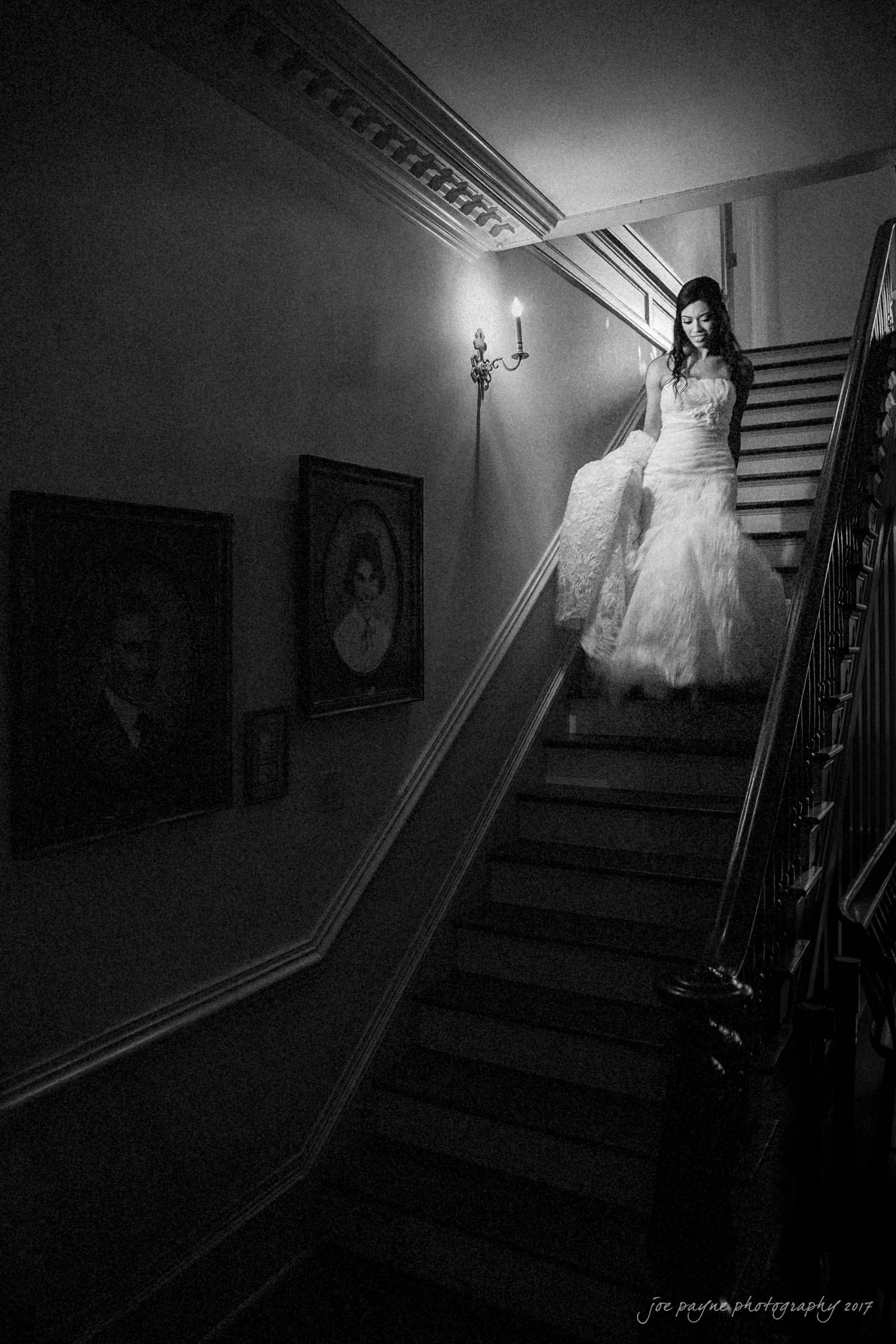 A bride in a long, flowing gown descends a dimly lit staircase. The walls are adorned with vintage portraits, and a single wall sconce casts a soft glow. The atmosphere is serene and timeless.