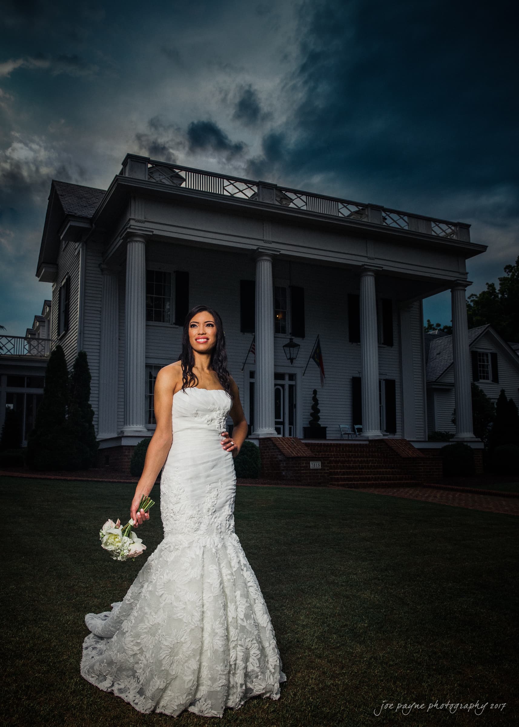 A woman in a strapless white wedding dress stands on a lawn in front of a large white mansion with columns. She holds a bouquet of flowers and gazes to the side under a dramatic cloudy sky.