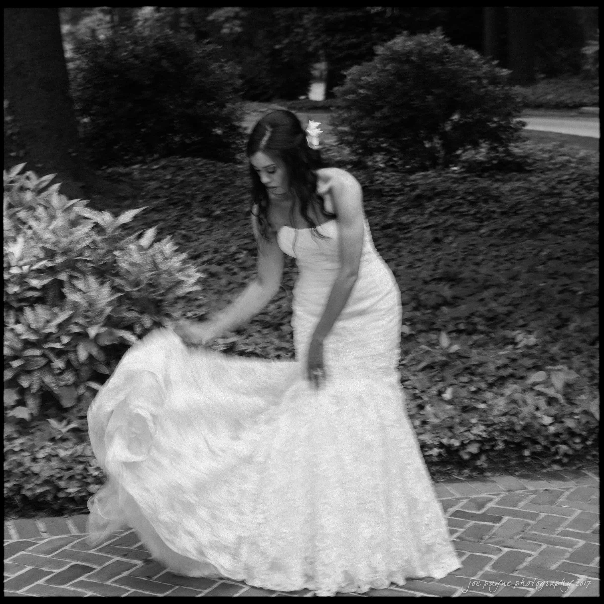 A woman in a white wedding dress stands on a brick path, holding her dress delicately. She is surrounded by lush greenery, including bushes and trees, creating a serene and elegant atmosphere. The image is in black and white.