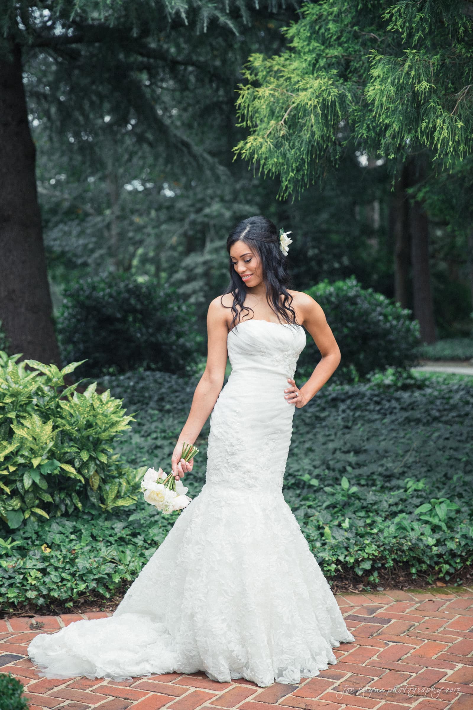 A bride in a strapless white wedding gown holding a bouquet stands on a brick path surrounded by lush greenery. She has long hair with a flower clip and is looking down with a serene expression.