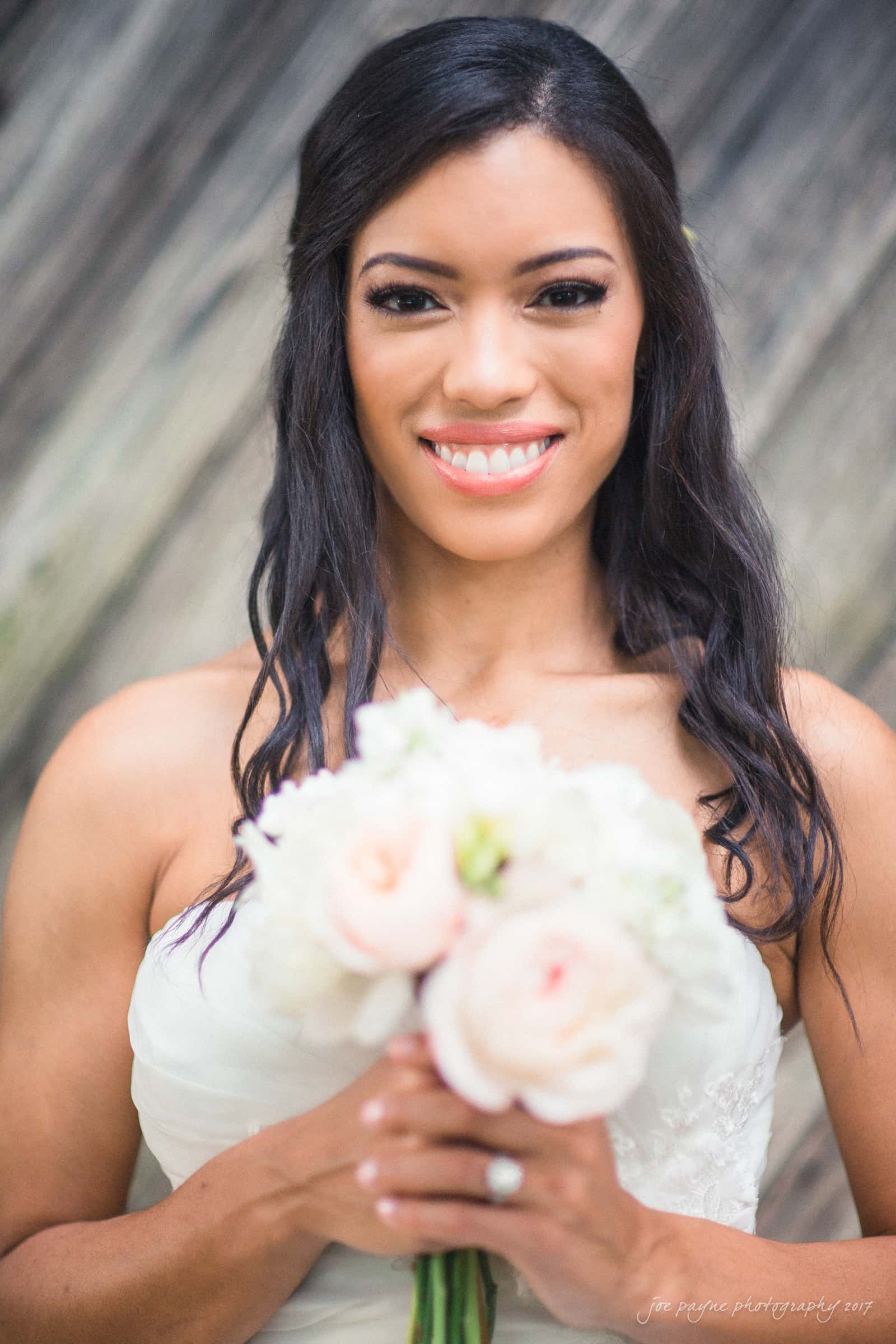 A bride in a strapless white dress holds a bouquet of light pink and white flowers. She has long, wavy dark hair and is smiling, standing against a textured wooden background.