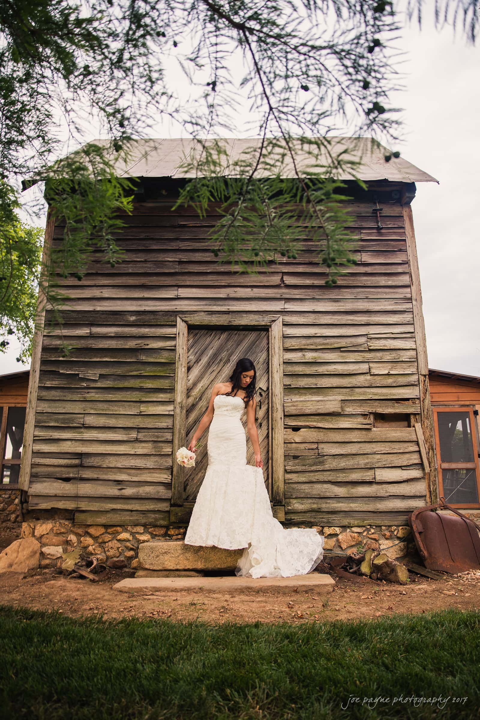 A bride in a white wedding dress stands on stone steps in front of a rustic wooden cabin. She holds a bouquet and is looking down. The scene is framed by green foliage, creating a natural, serene ambiance.