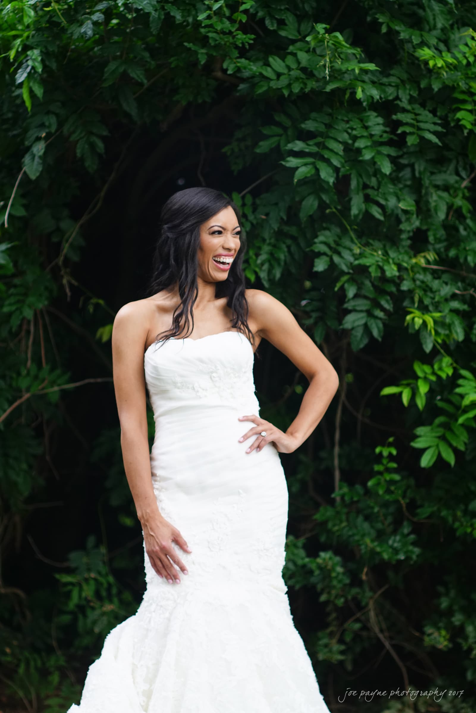 A woman in a strapless white wedding dress smiles and poses with a hand on her hip. She stands in front of lush green foliage, creating a natural, vibrant backdrop.